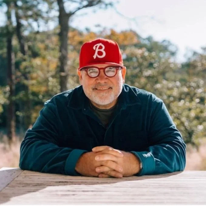 A smiling man with glasses and a beard, wearing a red baseball cap with a white 'B' and a dark jacket, sitting outdoors on a wooden surface with trees and foliage in the background.