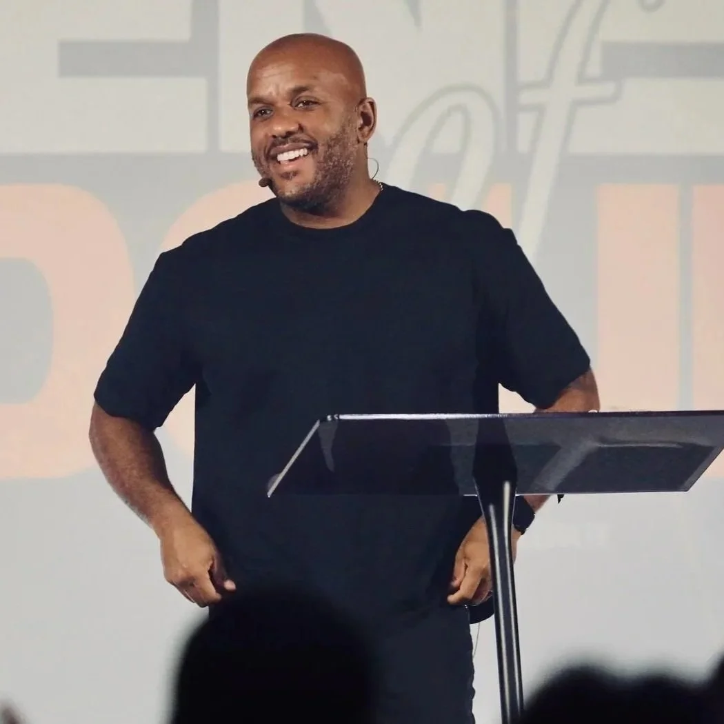 A man standing behind a music stand, wearing a black t-shirt, speaking or singing with a microphone headset, in front of a screen or wall with text in the background.