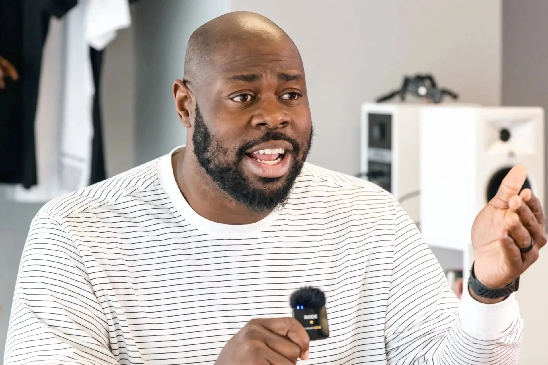A man with a beard and mustache speaking into a microphone with a tech or recording equipment in the background.