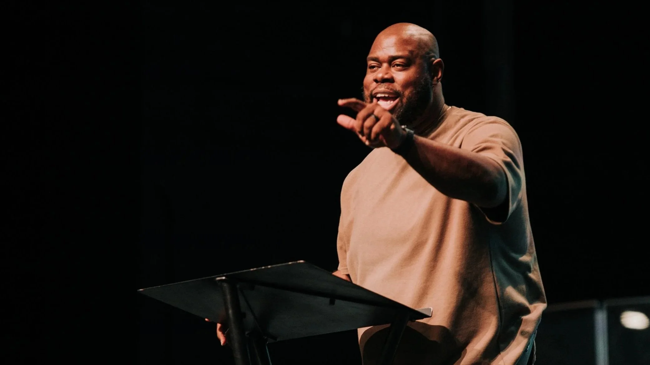 A man with a beard and bald head speaking on stage, gesturing with his right hand, standing behind a music stand, with a dark background.
