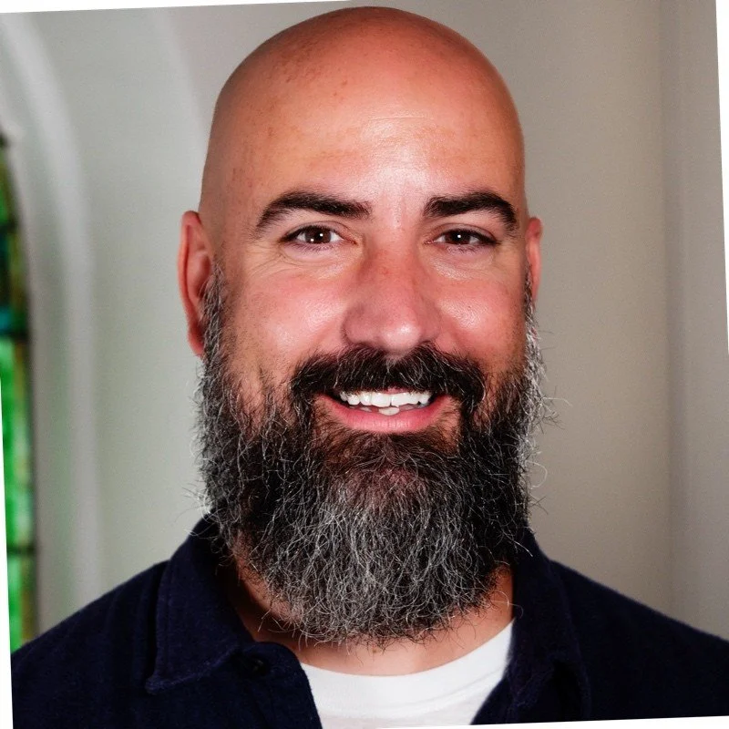 A smiling man with a bald head, full beard, and mustache, wearing a dark shirt and white undershirt, standing indoors.