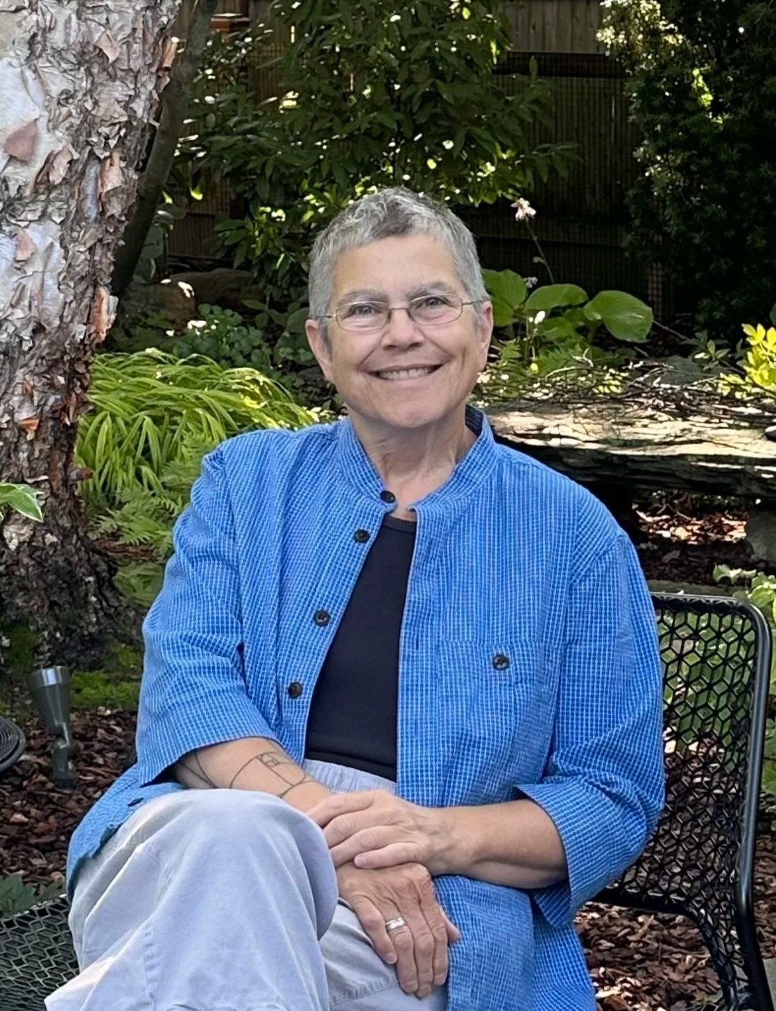 A woman with short gray hair wearing sunglasses, a gray tank top, blue shorts, and sandals standing next to a white pickup truck with a wooden flatbed. The truck has a sign that reads "Ground Flora" with a phone number, and is parked on grass with trees in the background.