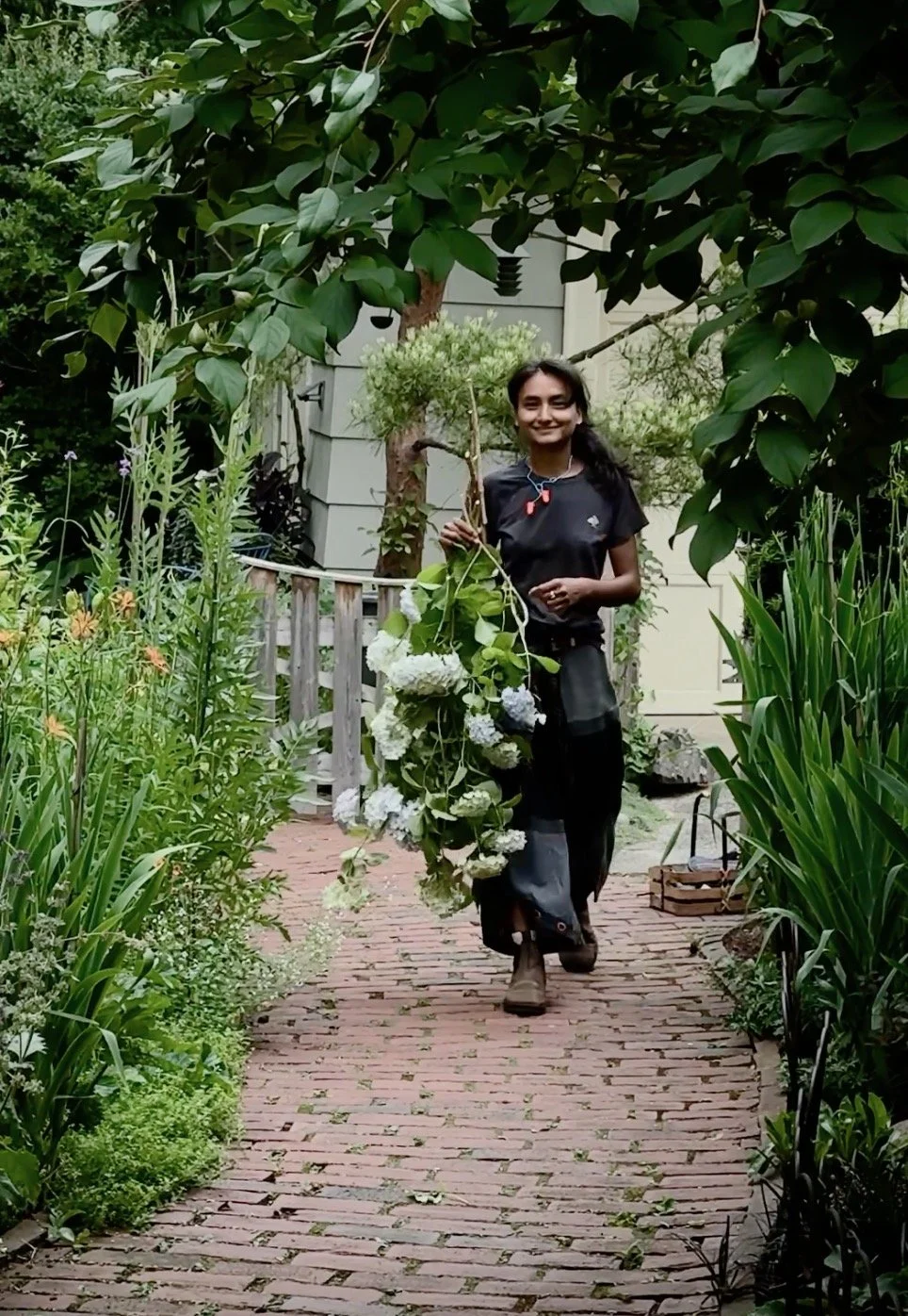 A person walking along a brick garden path, holding a branch with white flowers, surrounded by lush green plants and trees.