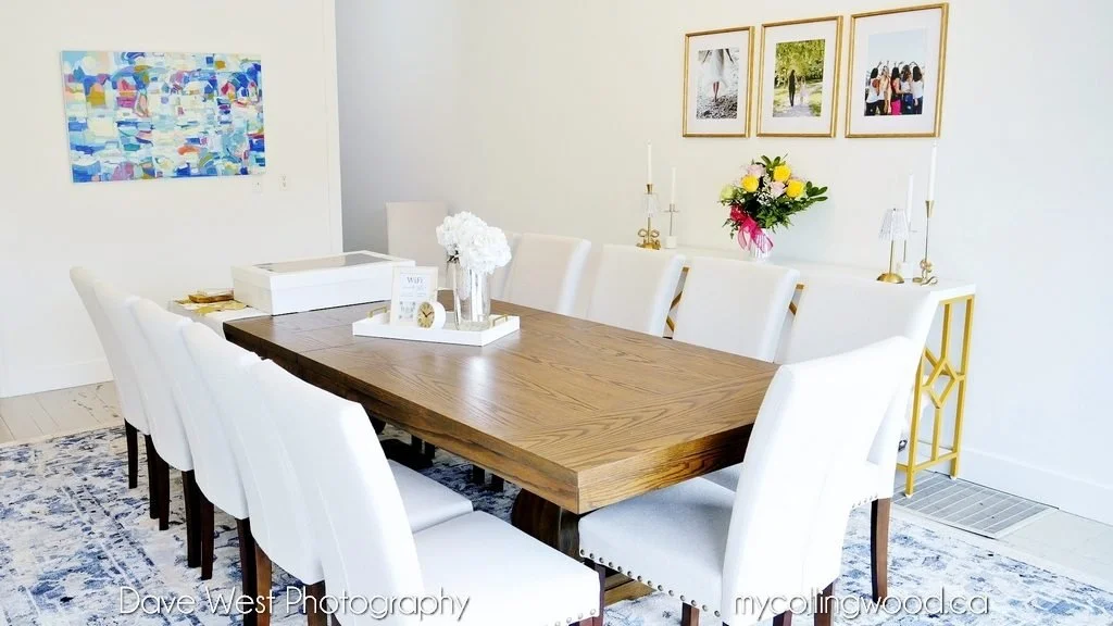 A dining room with a wooden table surrounded by eight white chairs, a broad blue and white rug, a sideboard with floral arrangement, framed photographs, and decorative candlesticks.