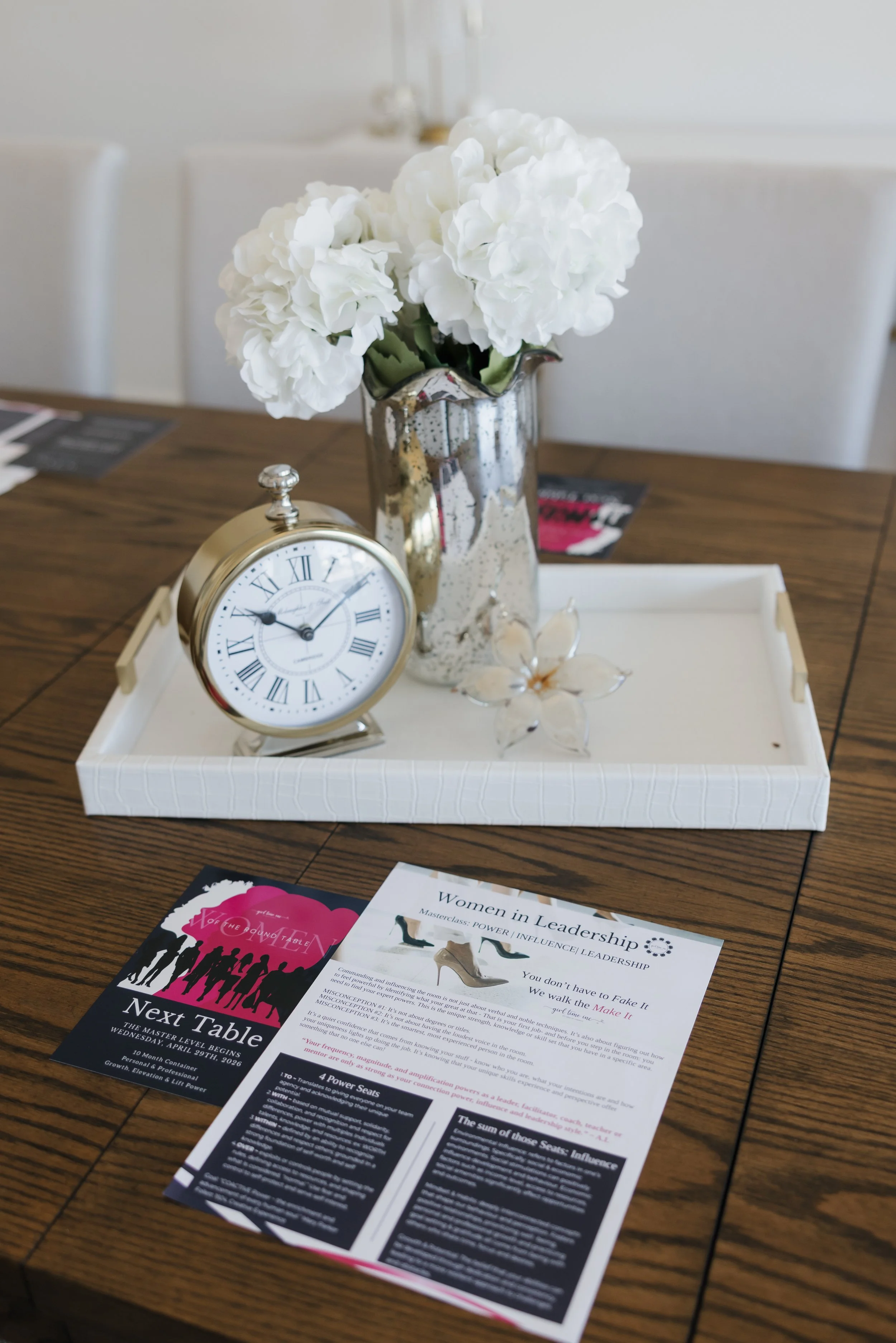 A table with a white tray holding a gold alarm clock, a clear glass flower ornament, and a pamphlet titled 'Women in Leadership' at a conference or seminar.
