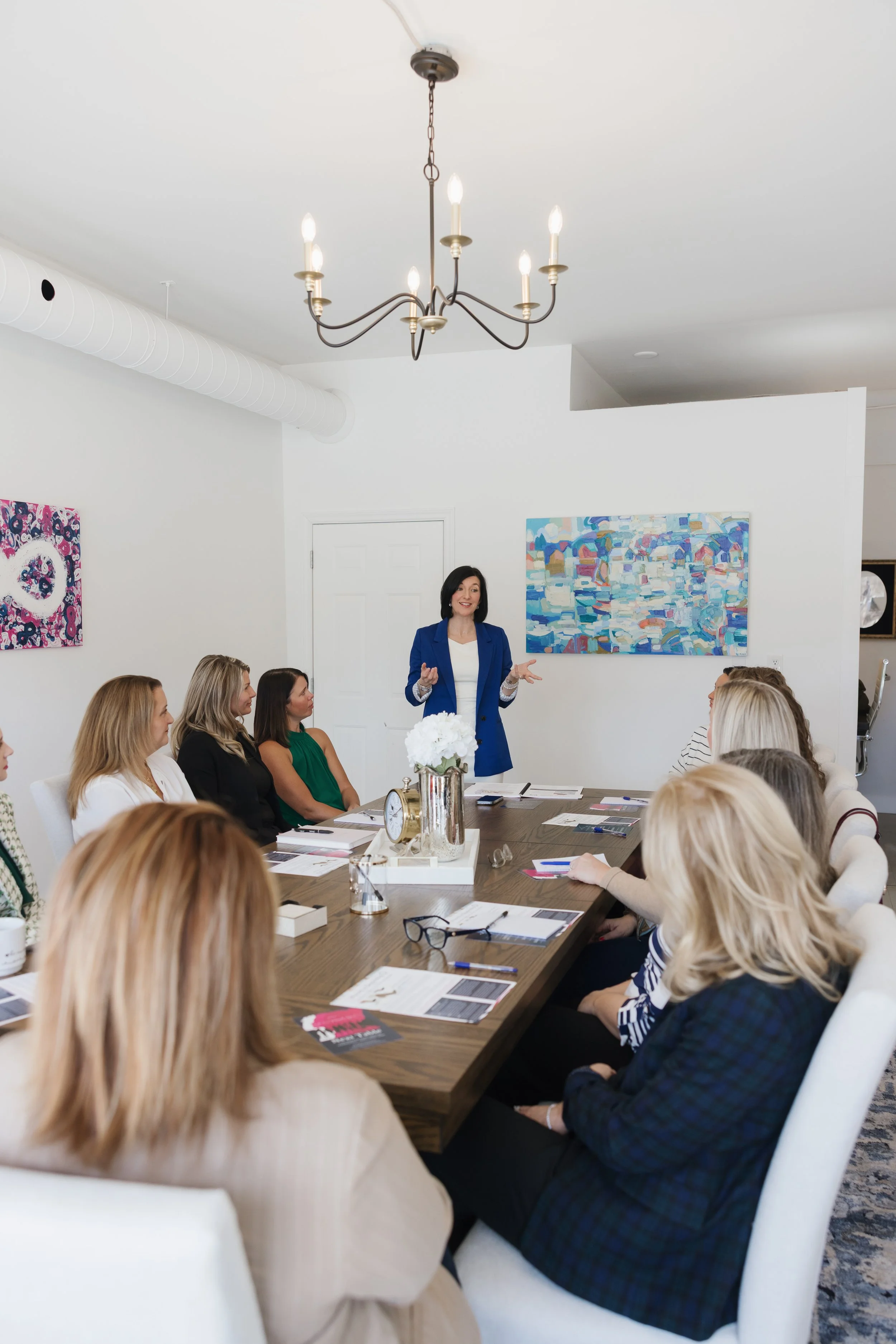 A woman in a blue blazer giving a presentation to a group of women seated around a large conference table in a bright room. There are abstract paintings on the white walls and a ceiling chandelier.