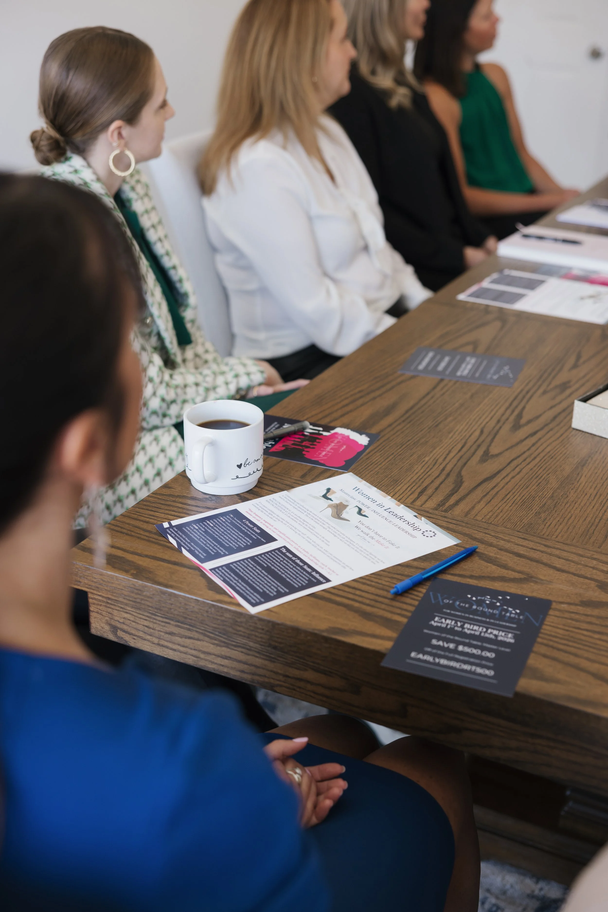 A group of women sitting at a meeting table, listening to a presentation. The table has brochures, flyers, a blue pen, and a coffee mug on it.