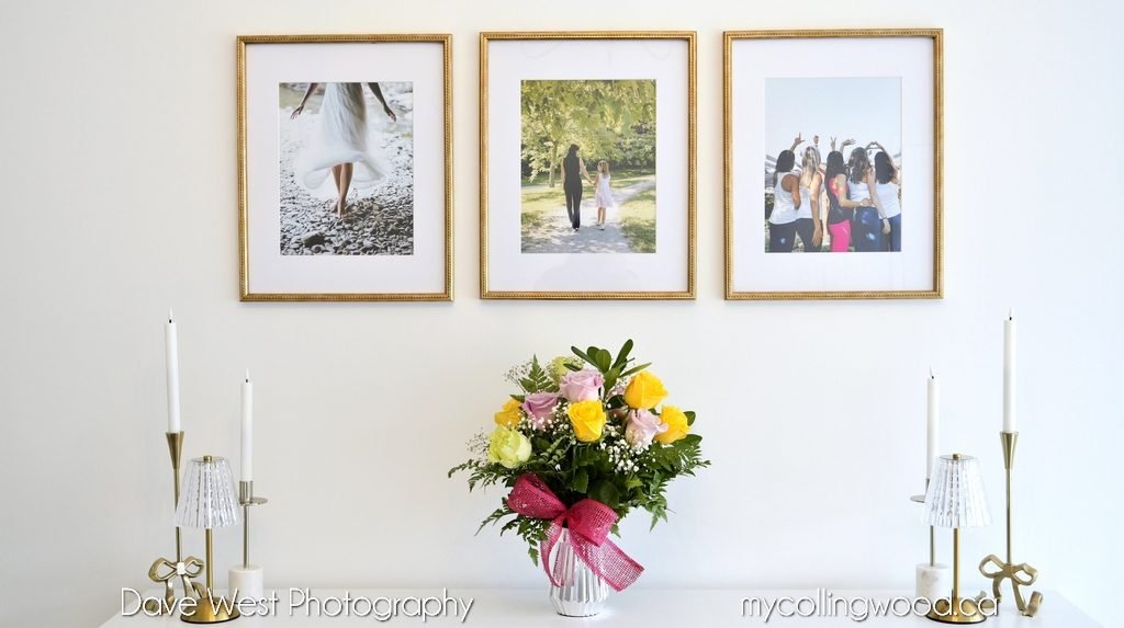 A white mantel with a flower arrangement in the center and four white candles in gold holders, with three framed family photos above.