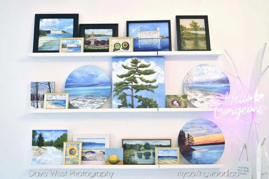 Display of various landscape paintings on white shelves, including seascapes, trees, and a sunset on a waterfront, with a neon sign reading "Hello Gorgeous" on the right side.