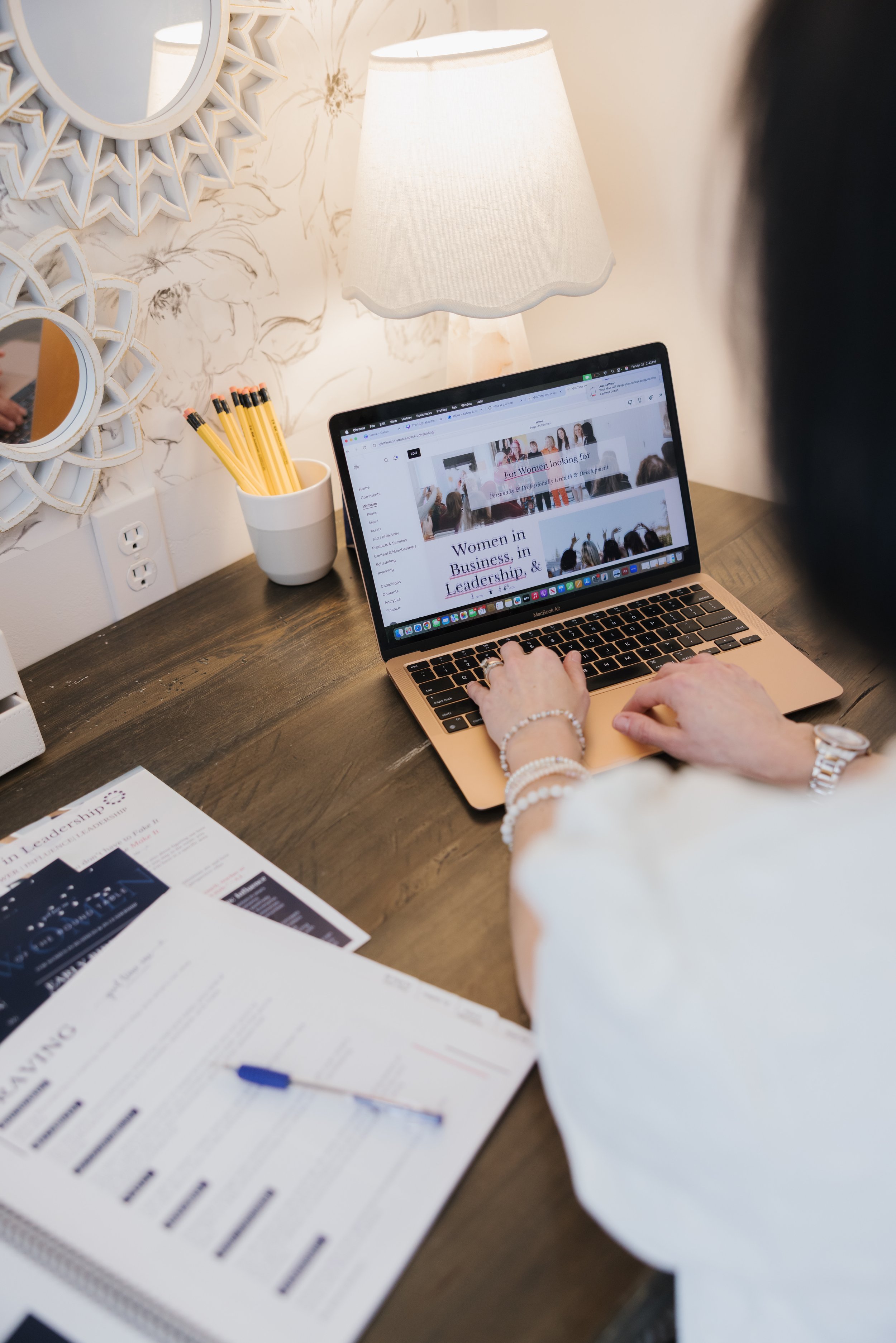 A person pointing at a laptop screen displaying a website about women in business, leadership, and growth, on a wooden desk with papers, a cup full of yellow pencils, a white lamp, and decorative mirrors on the wall.