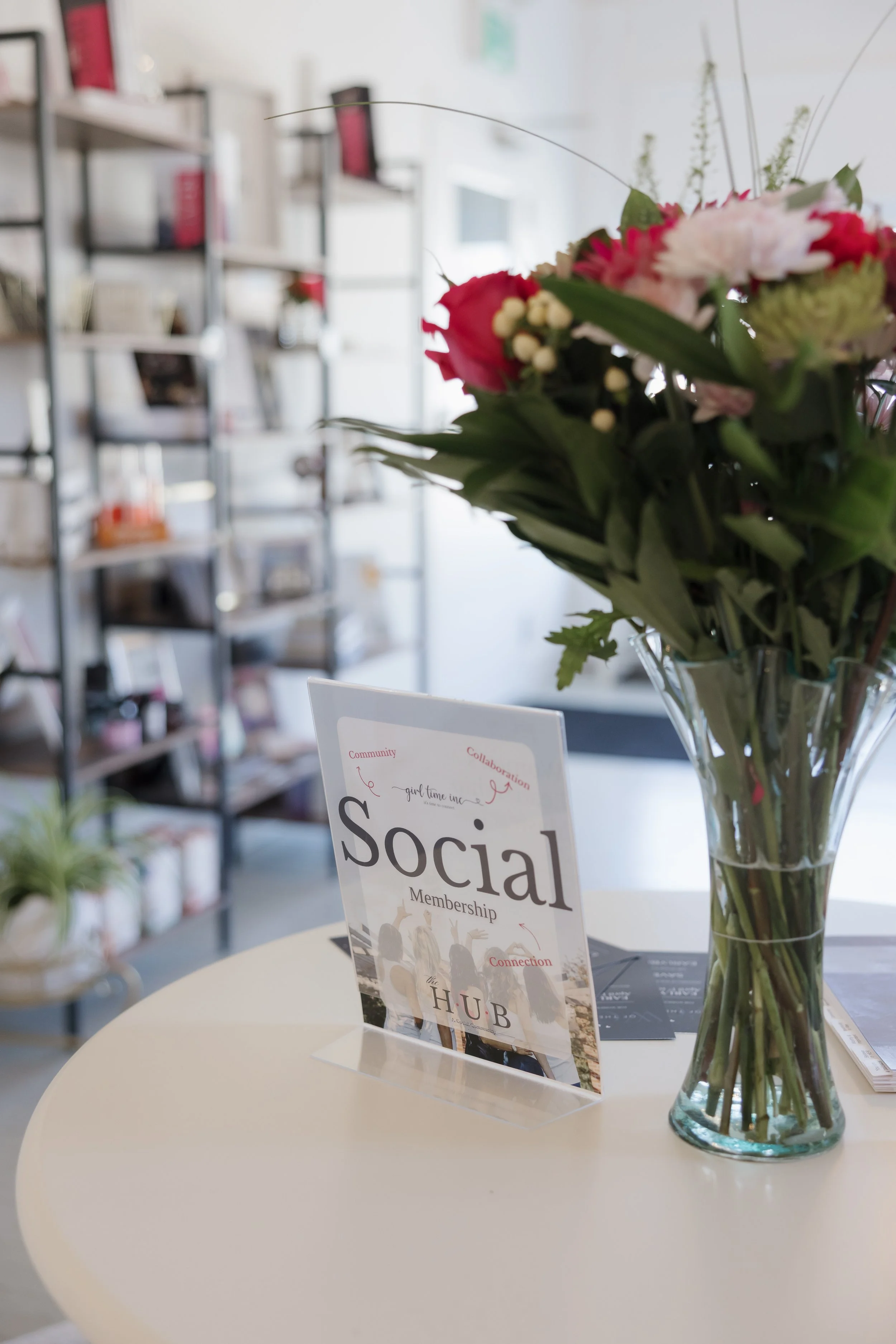 A clear glass vase filled with a bouquet of pink and white flowers on a white table, with a sign about social membership and connection in the foreground.