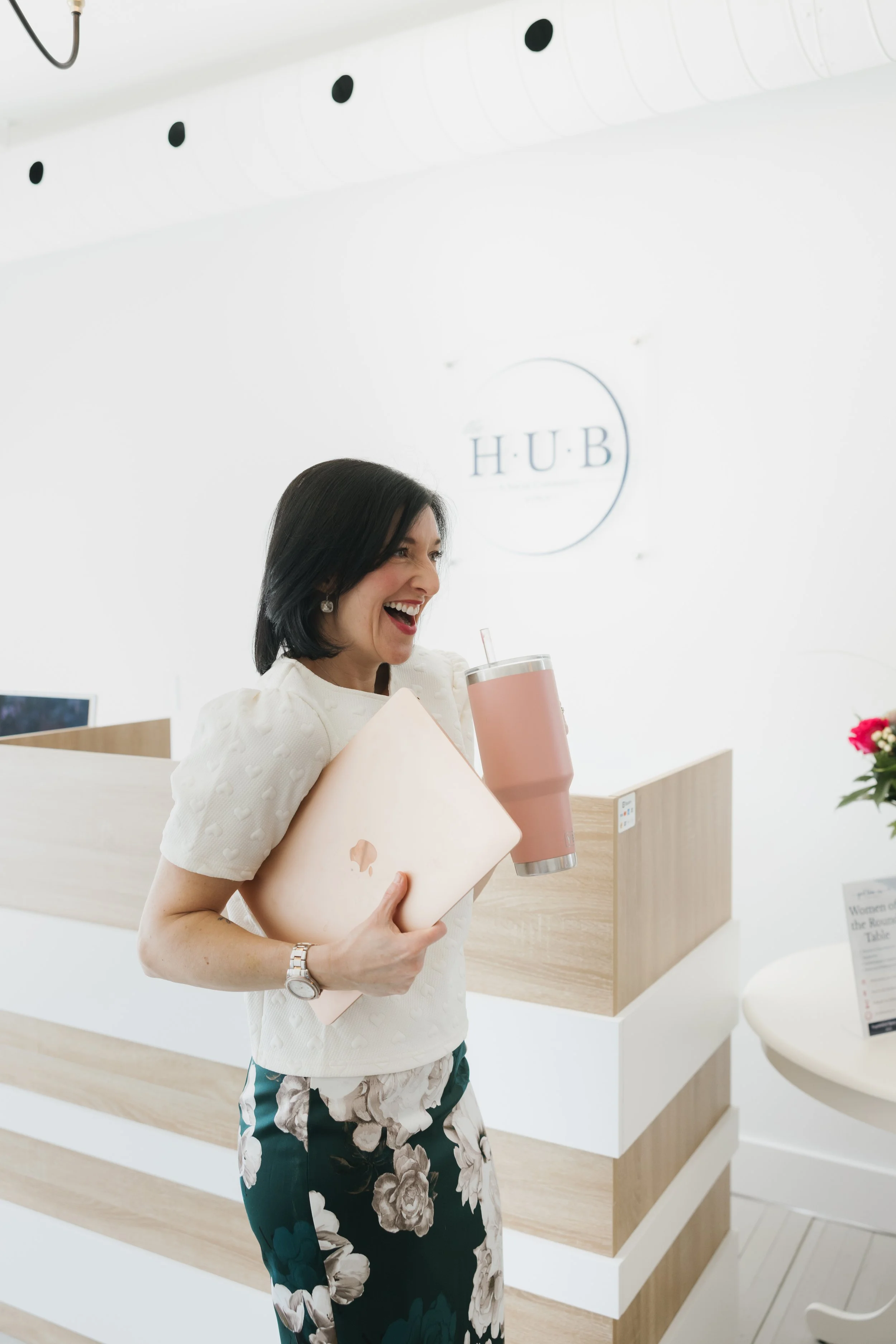 A woman with black hair and earrings is smiling and holding a closed rose gold laptop and a pink tumbler with a straw. She is in a bright, white space with a sign that says 'HUB' behind her and wooden reception desks.