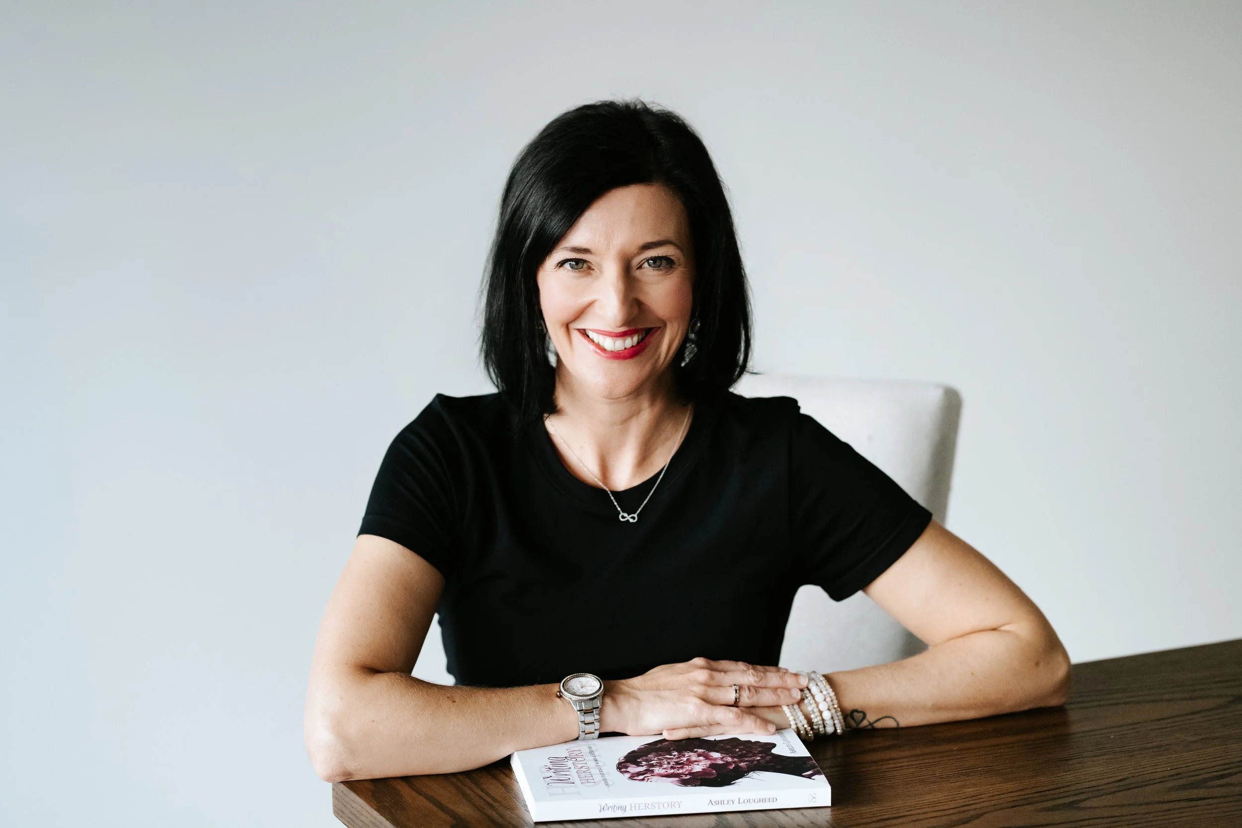 A woman with black hair and a bright smile sitting at a wooden table, wearing a black shirt, jewelry, and a watch, with a book titled 'Nothing' on it.