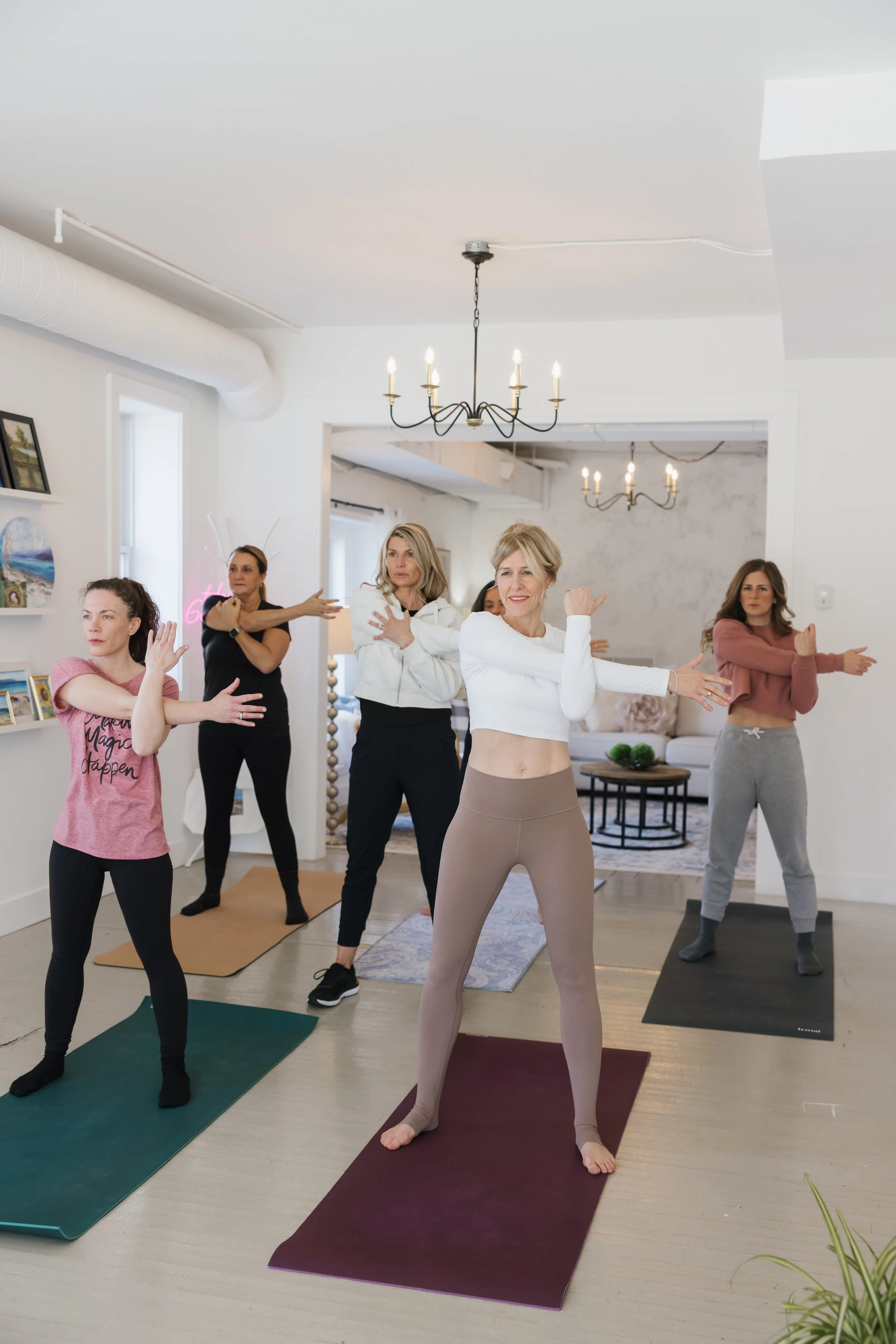 Group of women practicing yoga in a bright, airy room with white walls and black chandeliers.