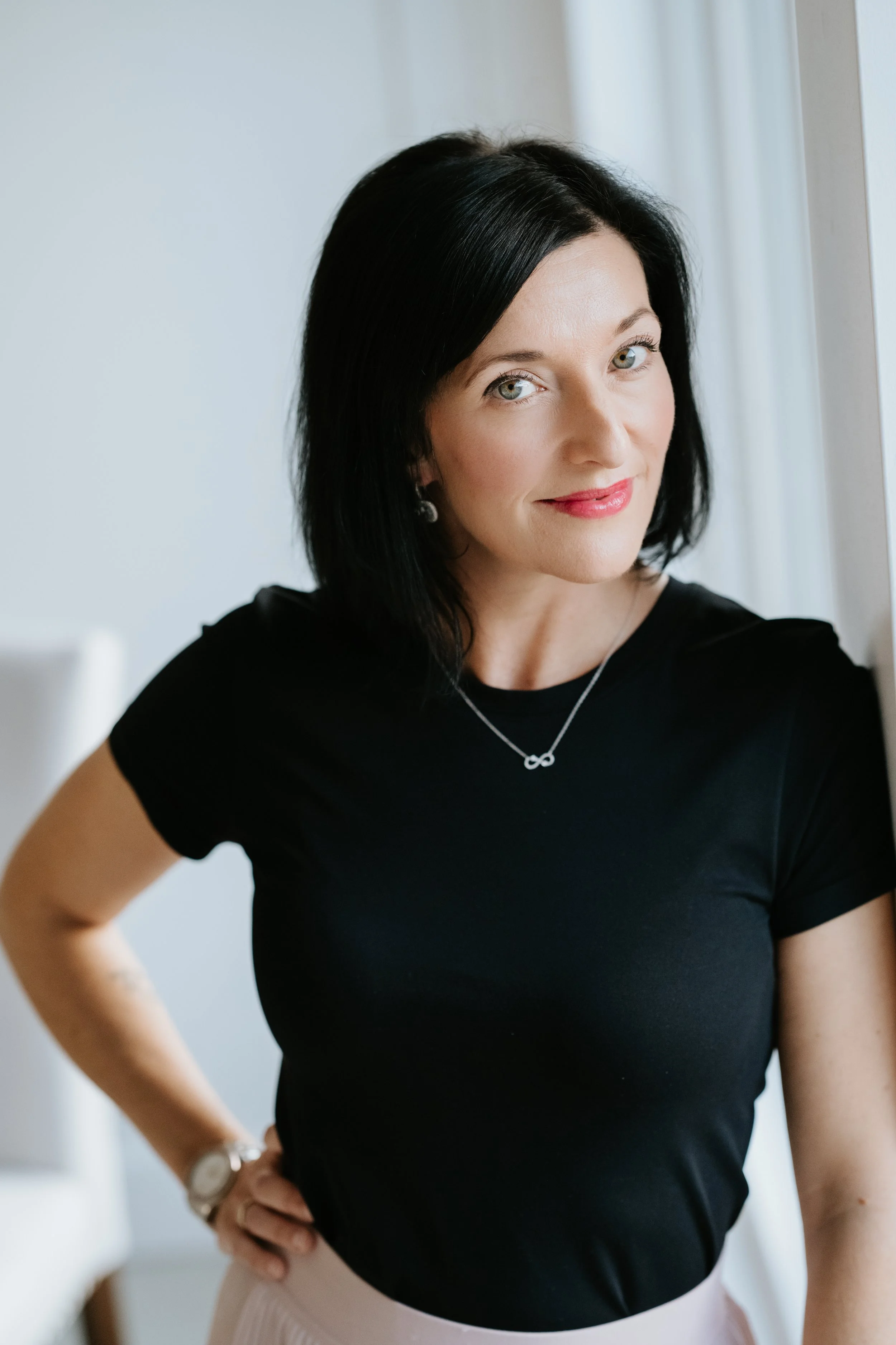 A woman with black hair, light skin, wearing a black shirt, standing near a window, smiling, with her right hand on her hip, wearing jewelry including a necklace, earrings, and a watch.