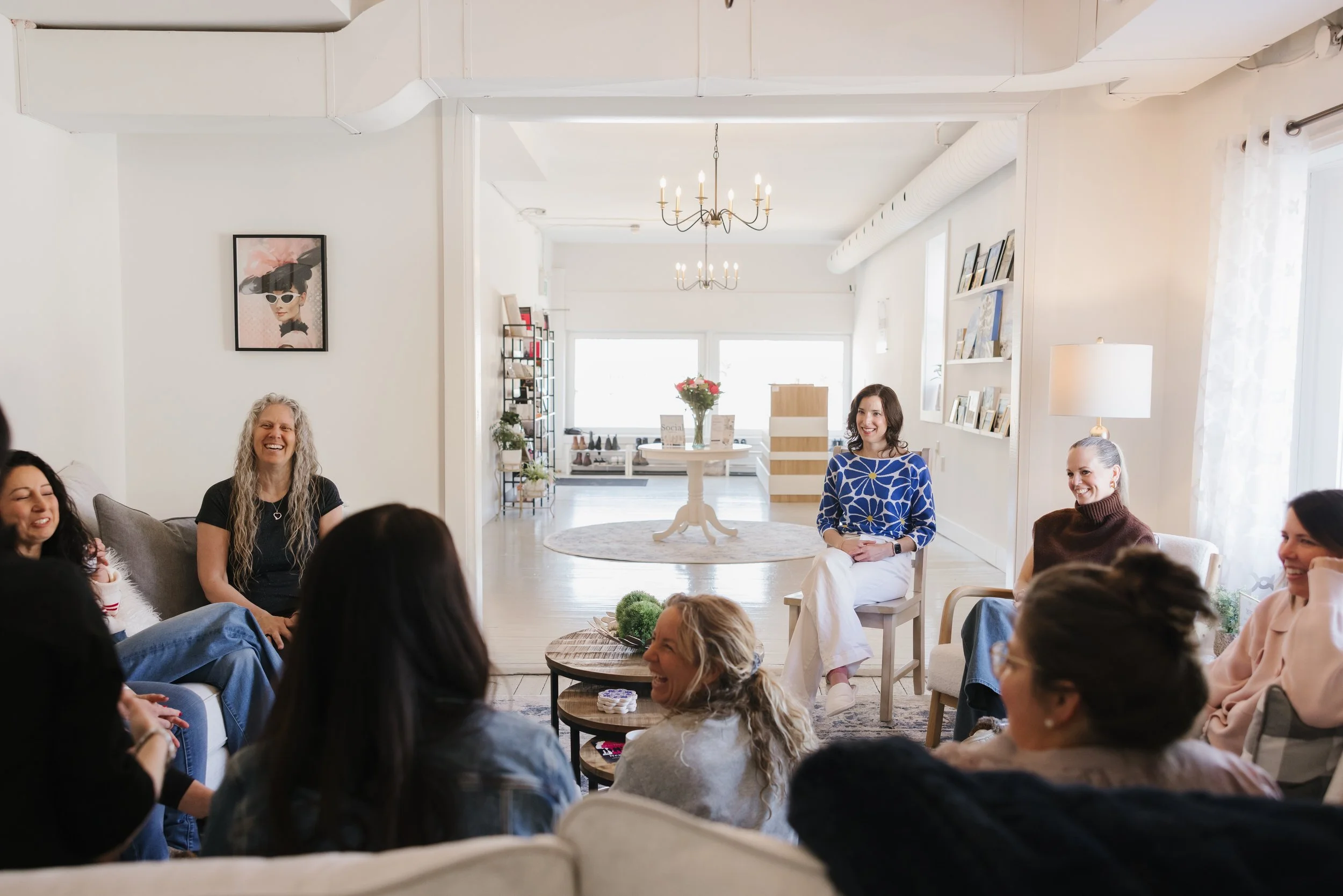 Group of women gathered in a bright, cozy room, smiling and engaging in conversation. The room is decorated with pictures, a chandelier, and a table with flowers.