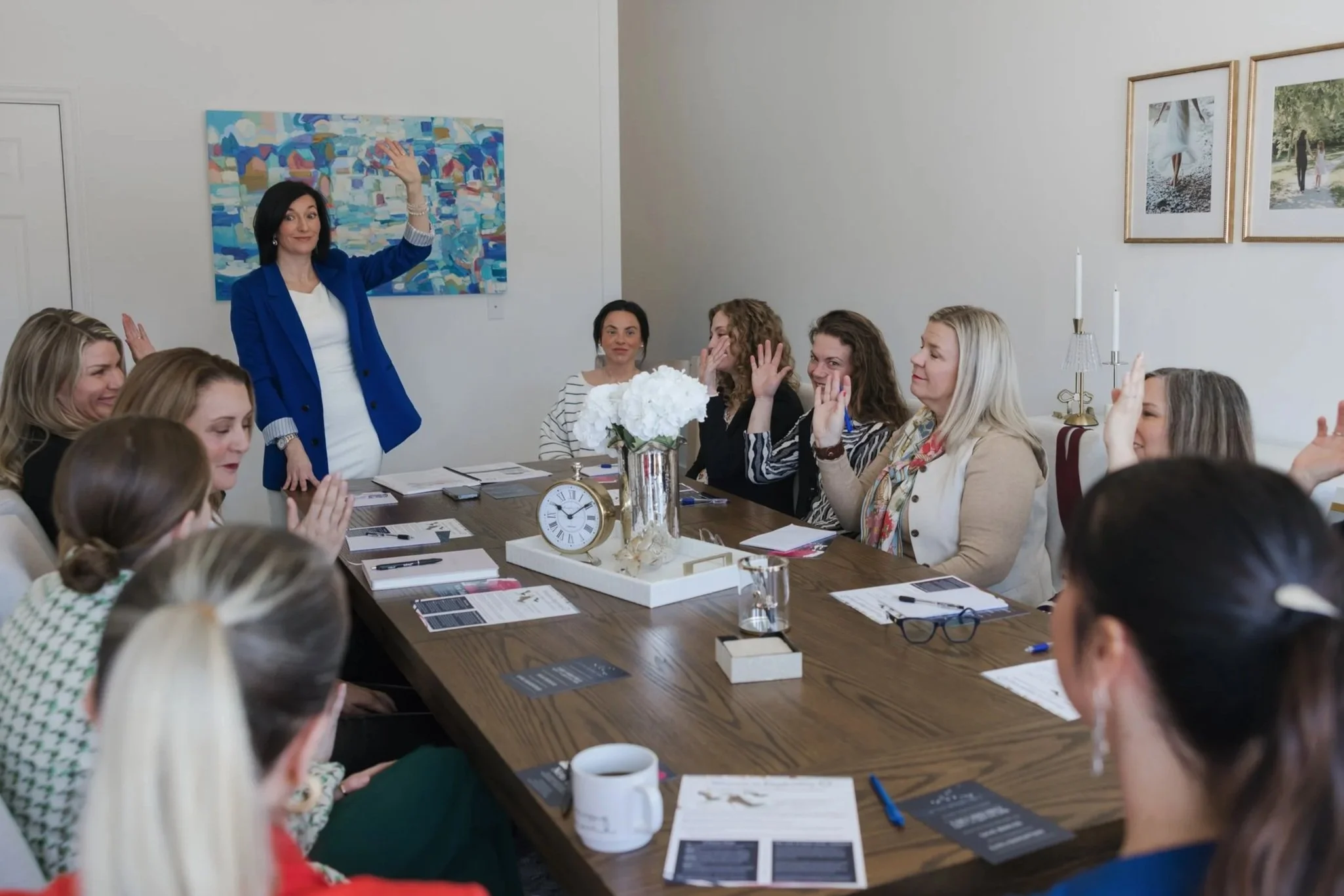 Group of ten women sitting and standing in a modern, stylish living room with artwork and decorative objects, smiling at the camera.