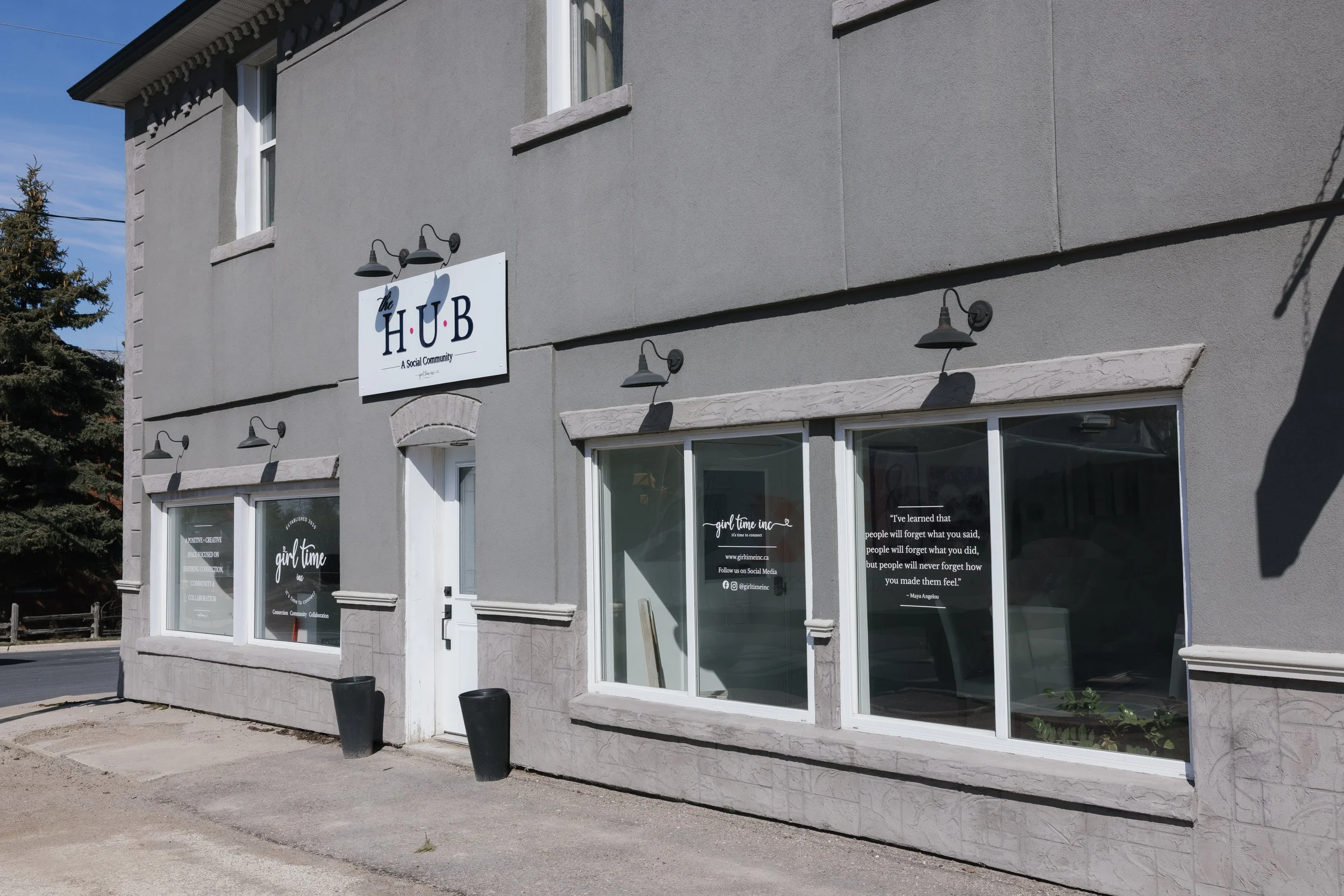 Exterior of a gray building with large windows, a sign that reads 'The H.U.B. A Social Community,' and smaller window decals with positive quotes and social media information.