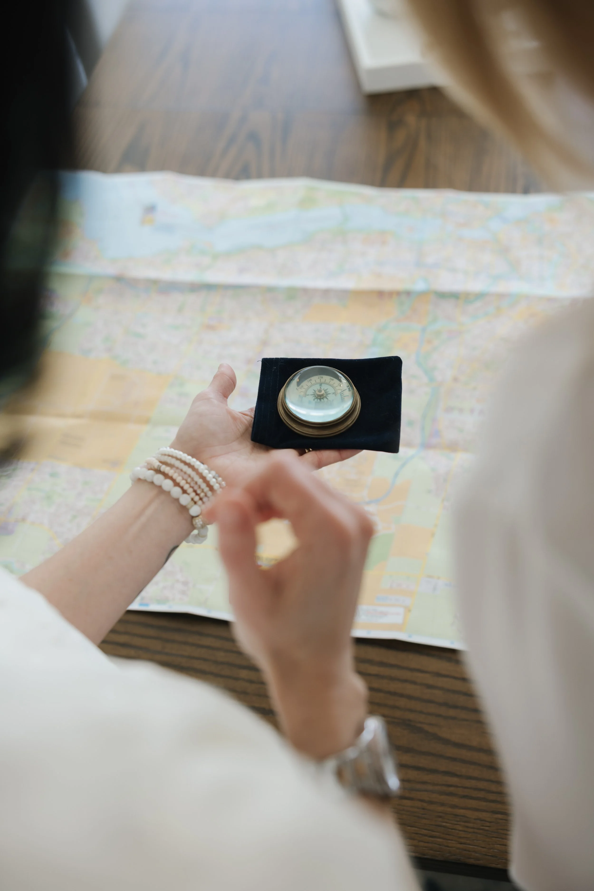 A person holding a compass in their hand over a map laid out on a wooden table.
