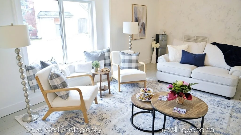Bright living room with large window, white sofa, two armchairs with plaid pillows, side tables, a round coffee table with a flower arrangement, and decorative items.