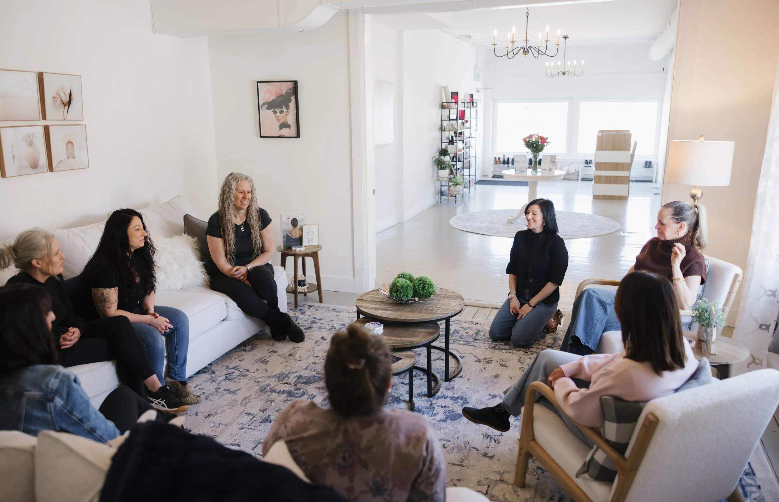 Group of women sitting and kneeling in a circle in a bright living room, engaged in conversation, with artwork on the walls and a coffee table with decorative moss in the center.