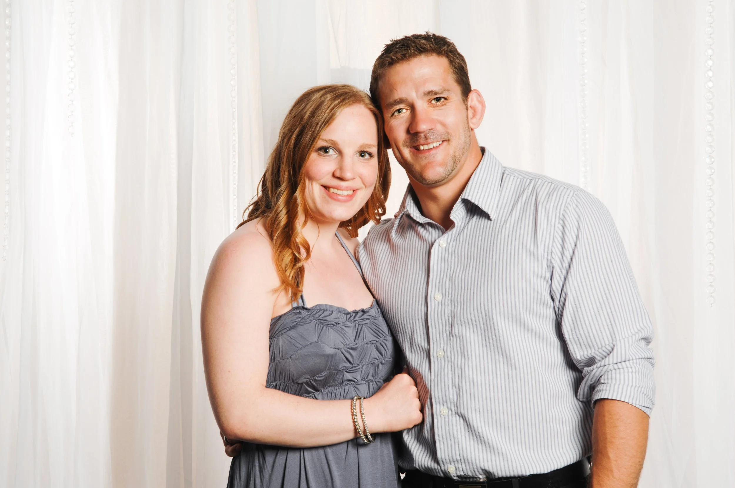 A couple standing together in front of a white curtain. The woman is wearing a strapless gray dress and the man is wearing a light-colored striped shirt.