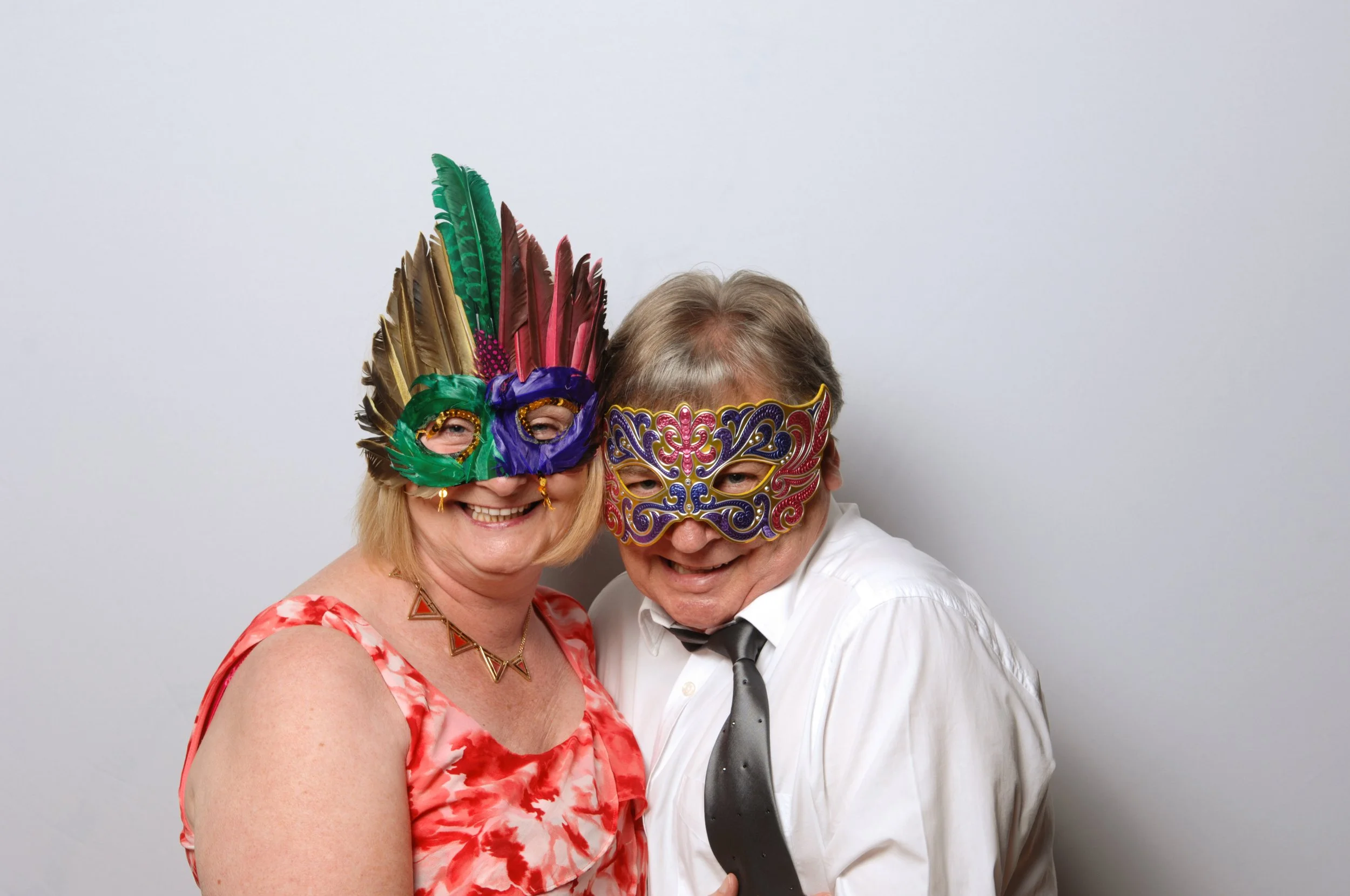 Two people wearing colorful masquerade masks. The person on the left has a mask with feathers in green, red, and gold, wearing a floral dress. The person on the right has a decorative red, blue, and gold mask, wearing a white shirt and dark tie.