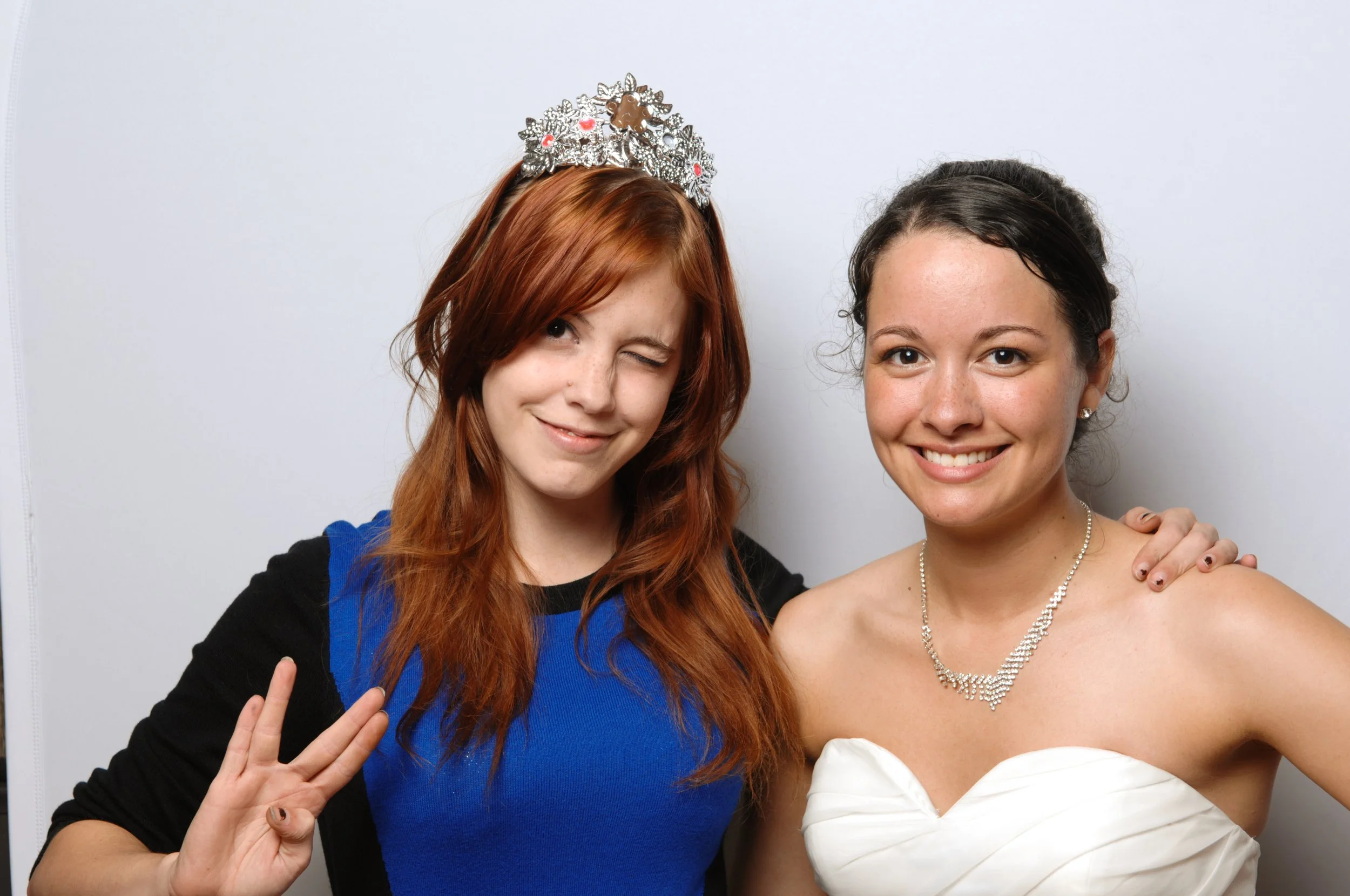 Two women posing together, one wearing a tiara and flashing a hand gesture, the other in a white dress smiling.