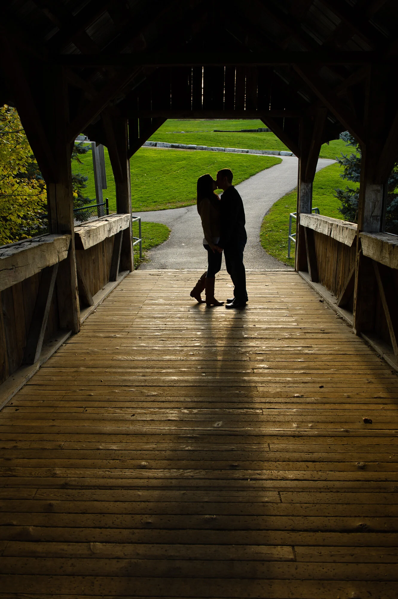Silhouette of a couple standing in a wooden covered bridge with a sunlit path outside.