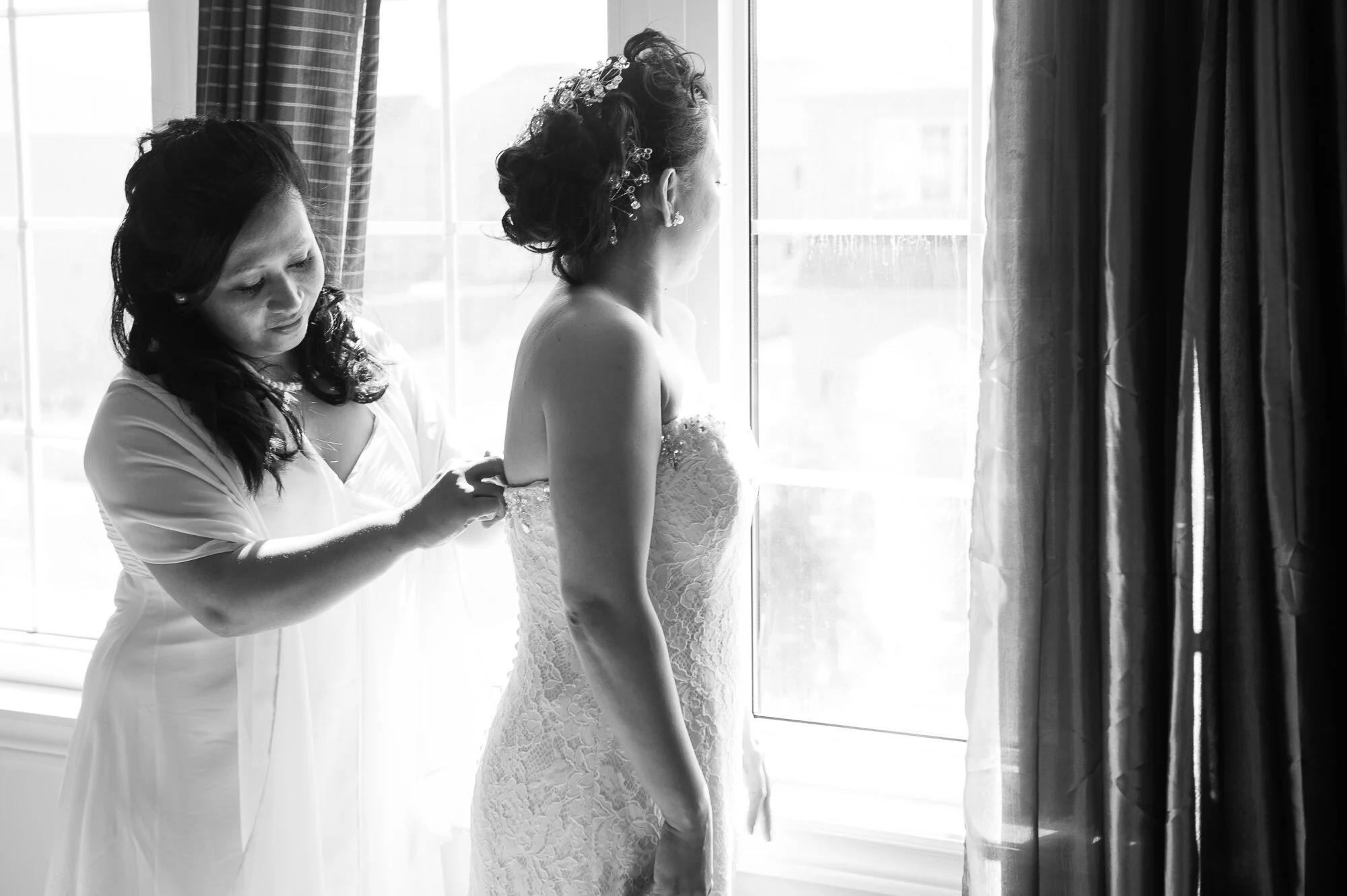 Woman in wedding dress being assisted by another woman near a window, black and white image.
