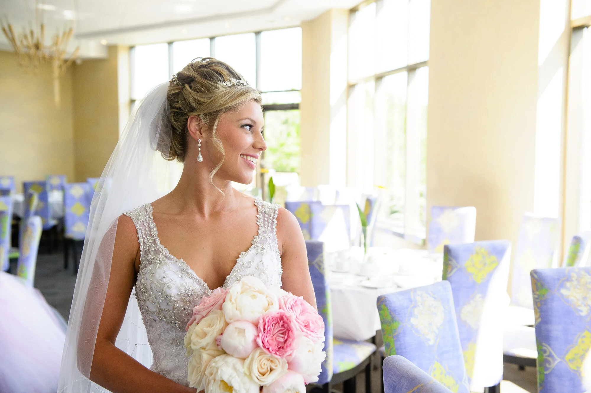 Bride in a wedding dress holding a bouquet of pink and white flowers, smiling, with a dining area in the background.
