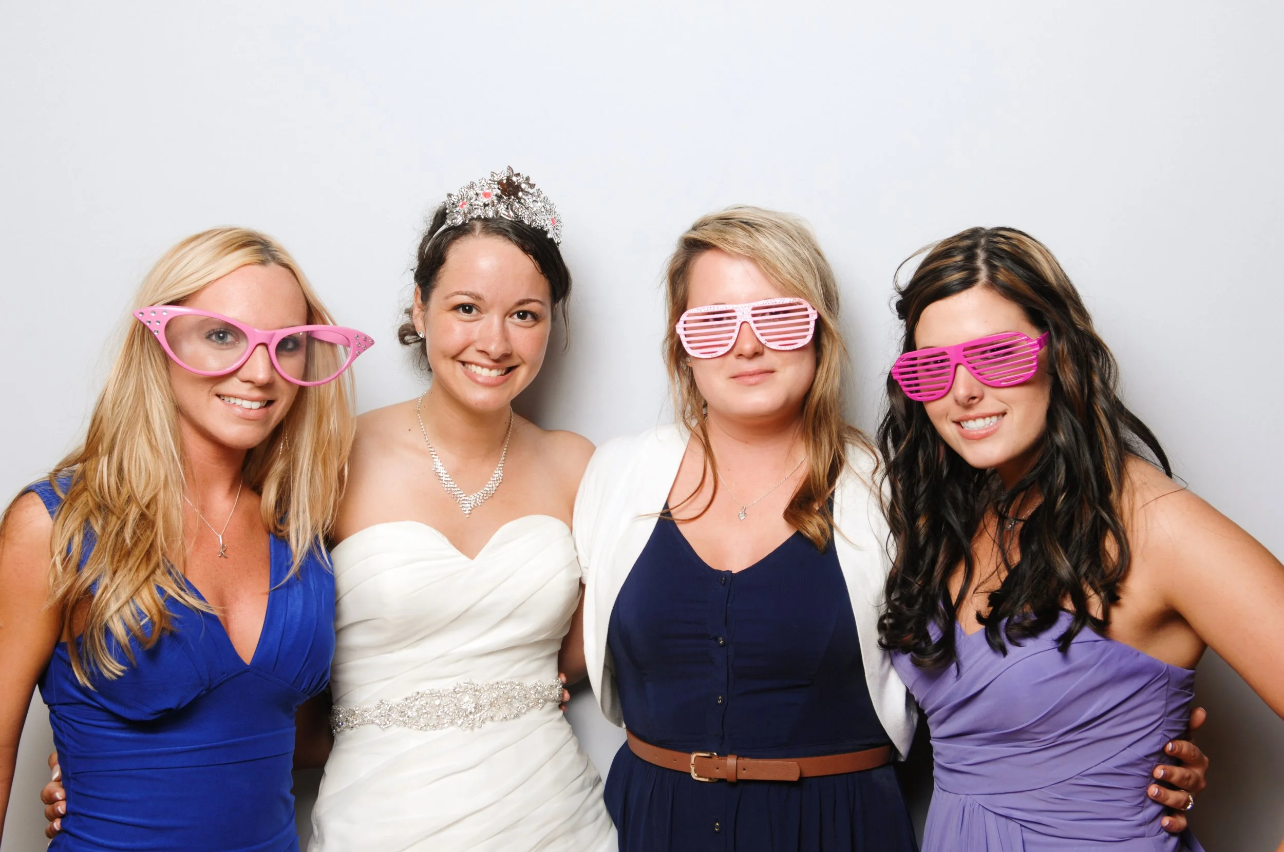 Four women posing for a fun photo, two wearing festive glasses, one in a wedding gown, and others in formal dresses.