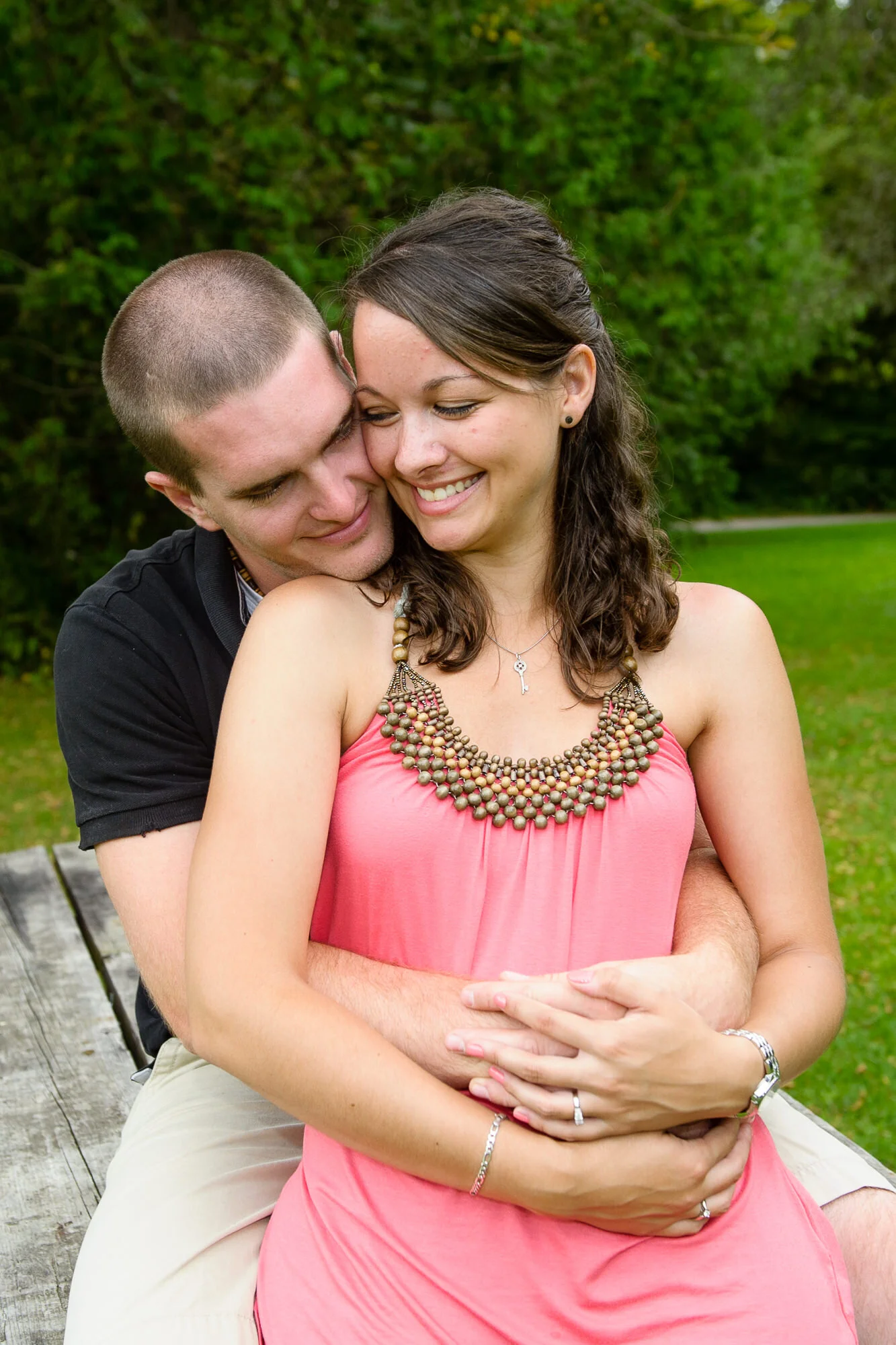 A couple sitting on a wooden bench. The woman is wearing a coral dress with a beaded neckline, and the man is wearing a black shirt. They are embracing and smiling, surrounded by greenery.