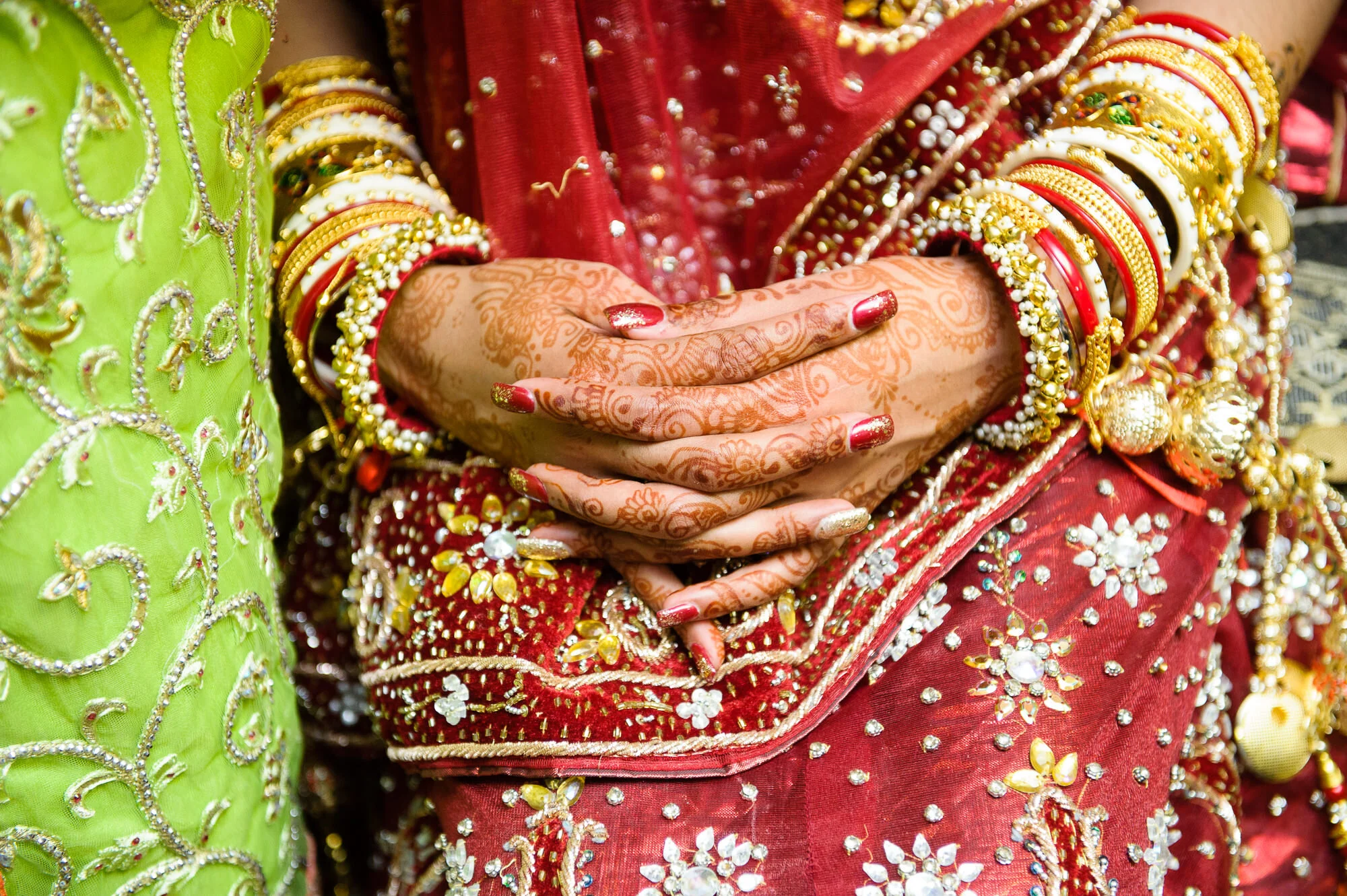 Close-up of hands with henna and bangles, wearing traditional red and green embroidered attire.