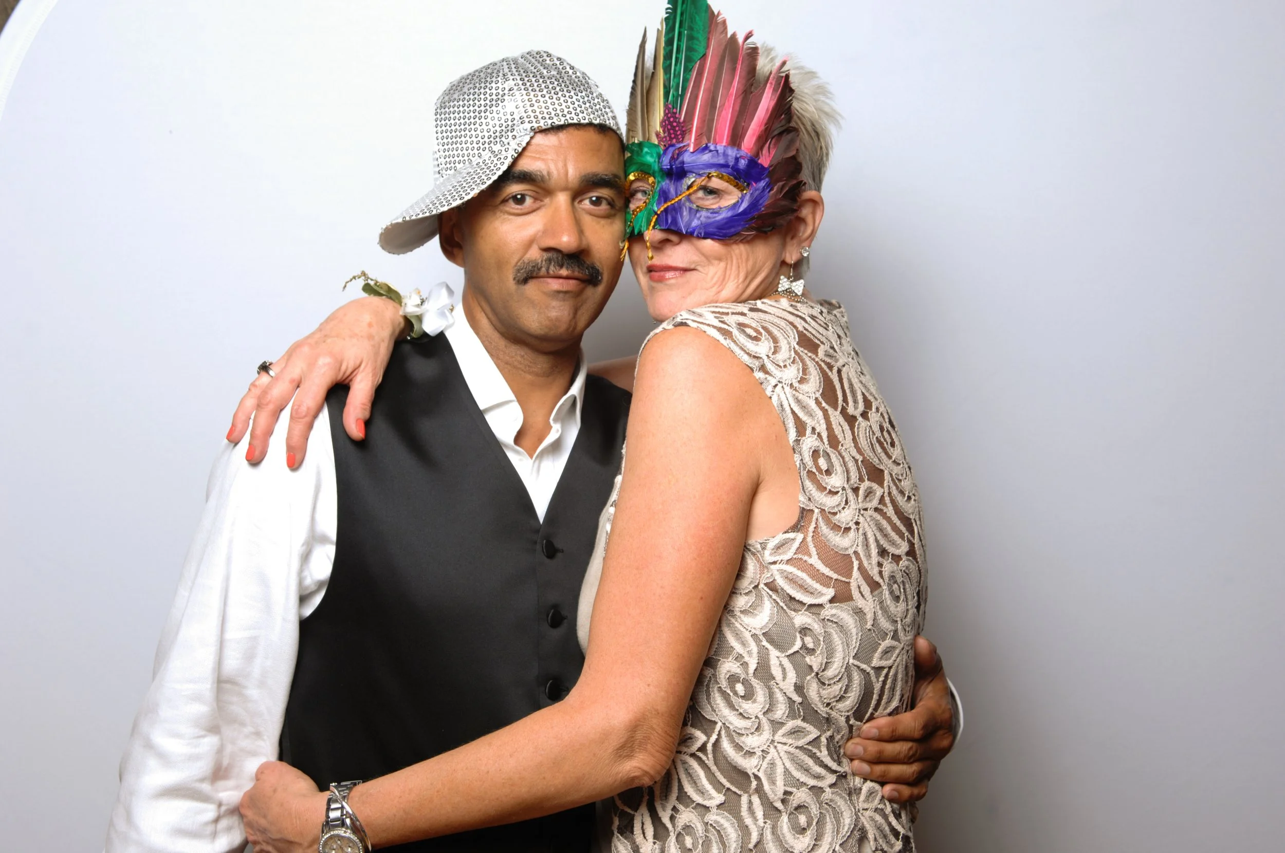 A couple posing for a photo, with the man wearing a white shirt, black vest, and a sequined cap, and the woman wearing a lace dress and colorful feathered mask.