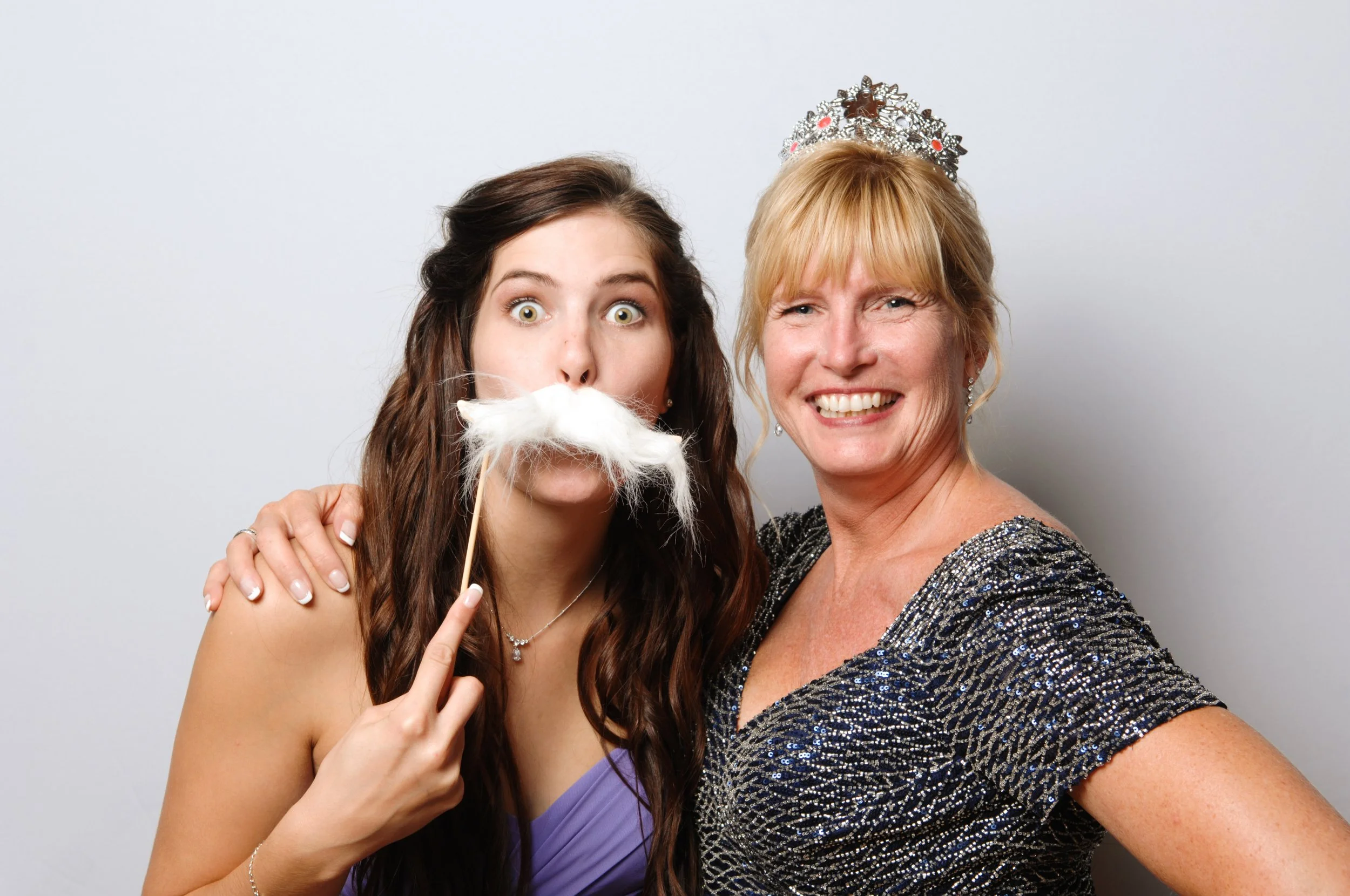 Two women posing for a playful photo, one holding a fake white mustache on a stick, wearing formal dresses and one with a tiara.