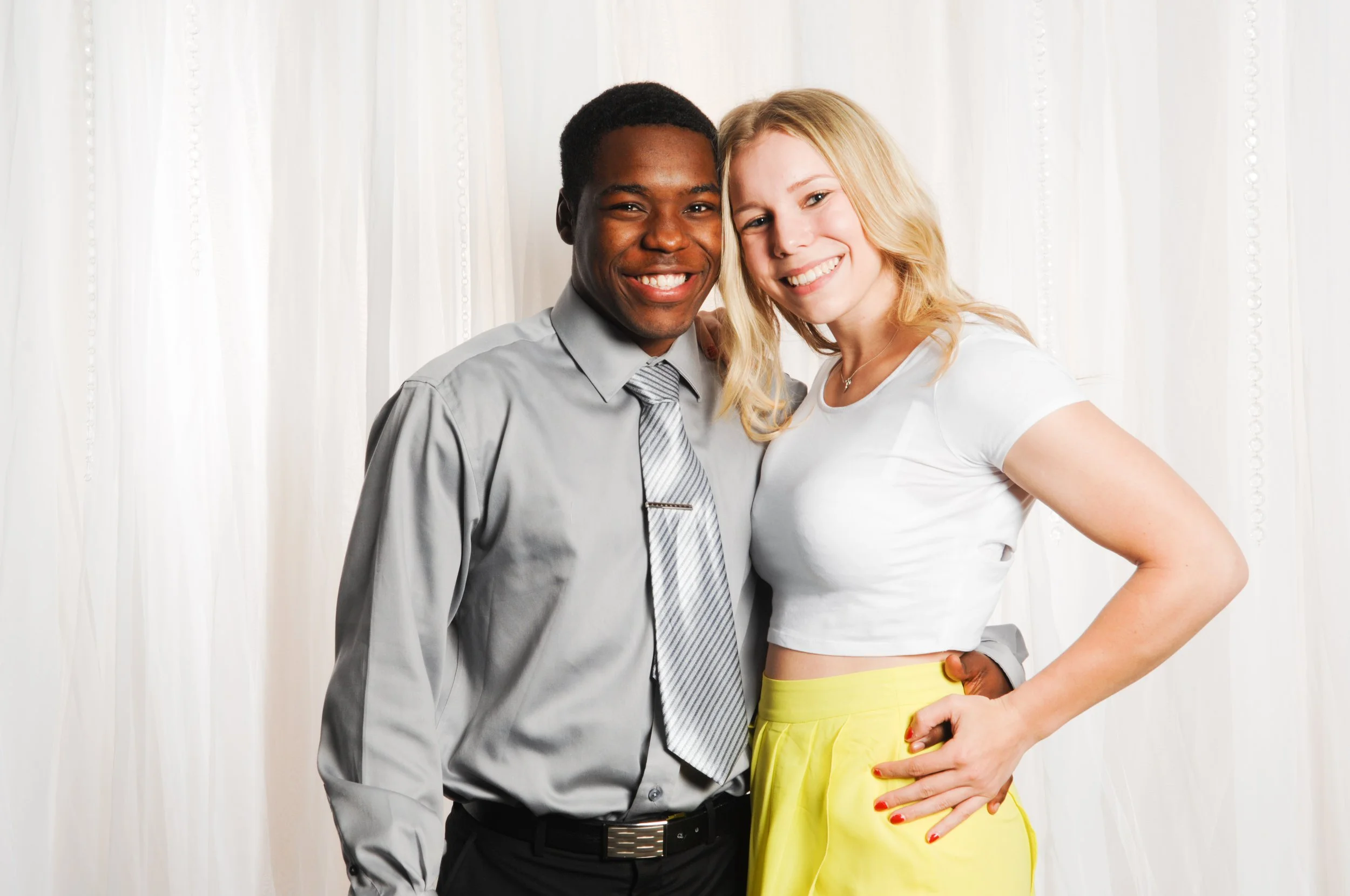 Two people smiling in front of a white curtain; one in a gray shirt and tie, the other in a white top and yellow skirt.