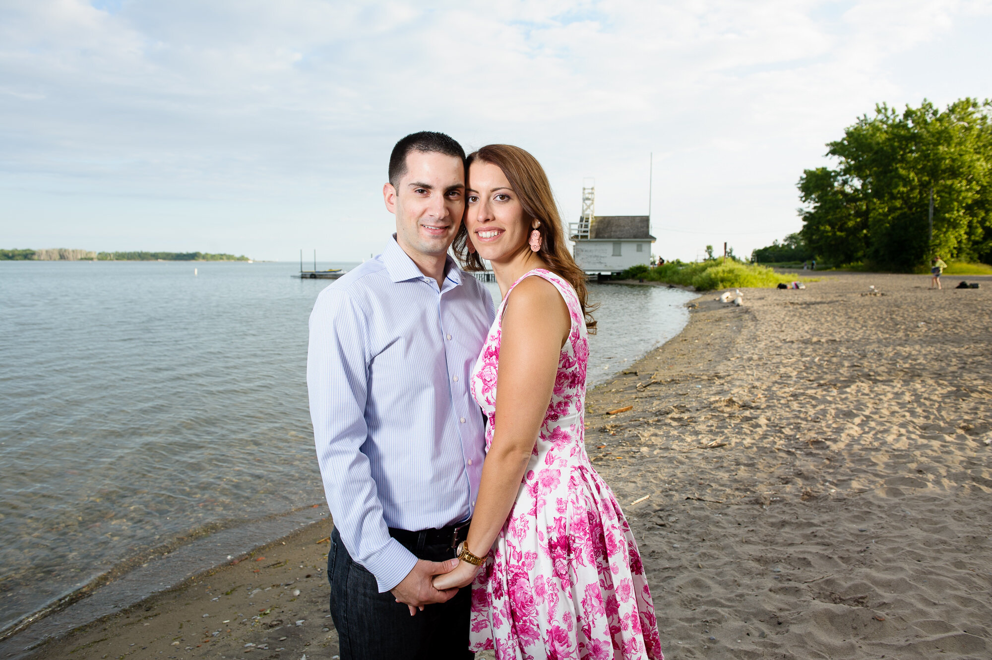 Couple posing on a sandy beach by calm water and green trees.