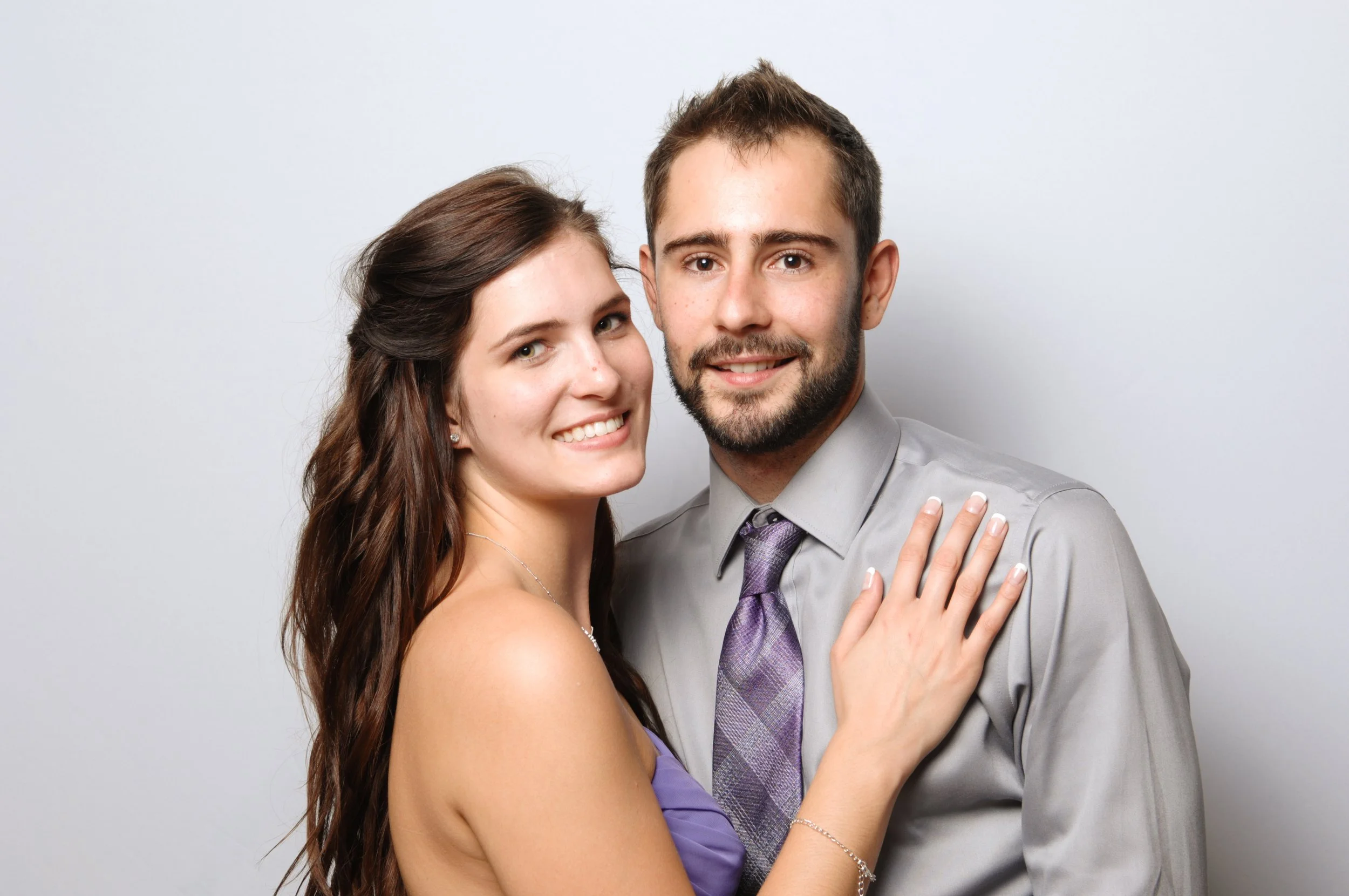 A smiling couple posing together, with the woman wearing a purple dress and the man in a gray shirt and purple tie.