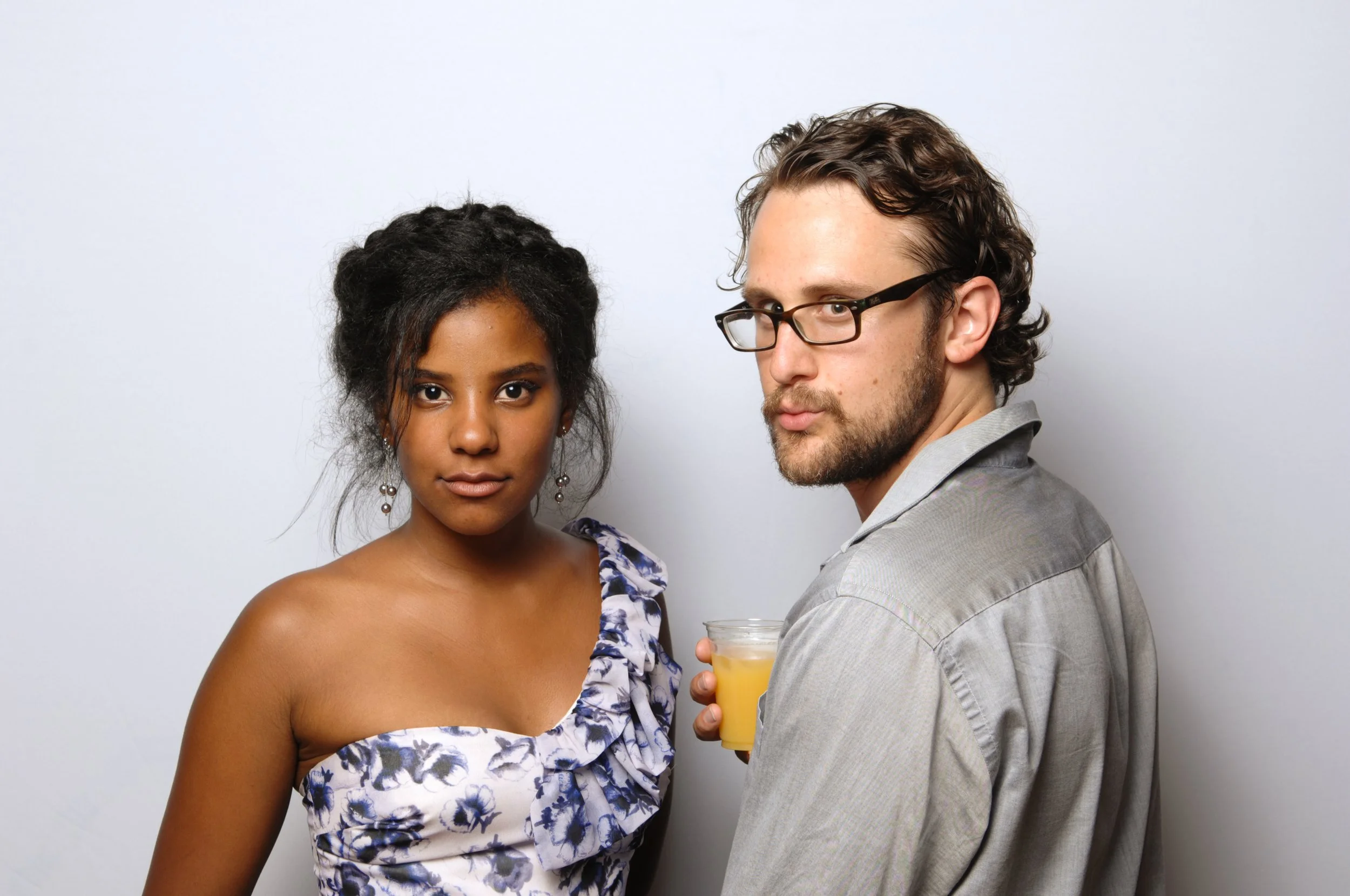 A woman in a floral dress and a man wearing glasses holding a drink, posing against a plain background.