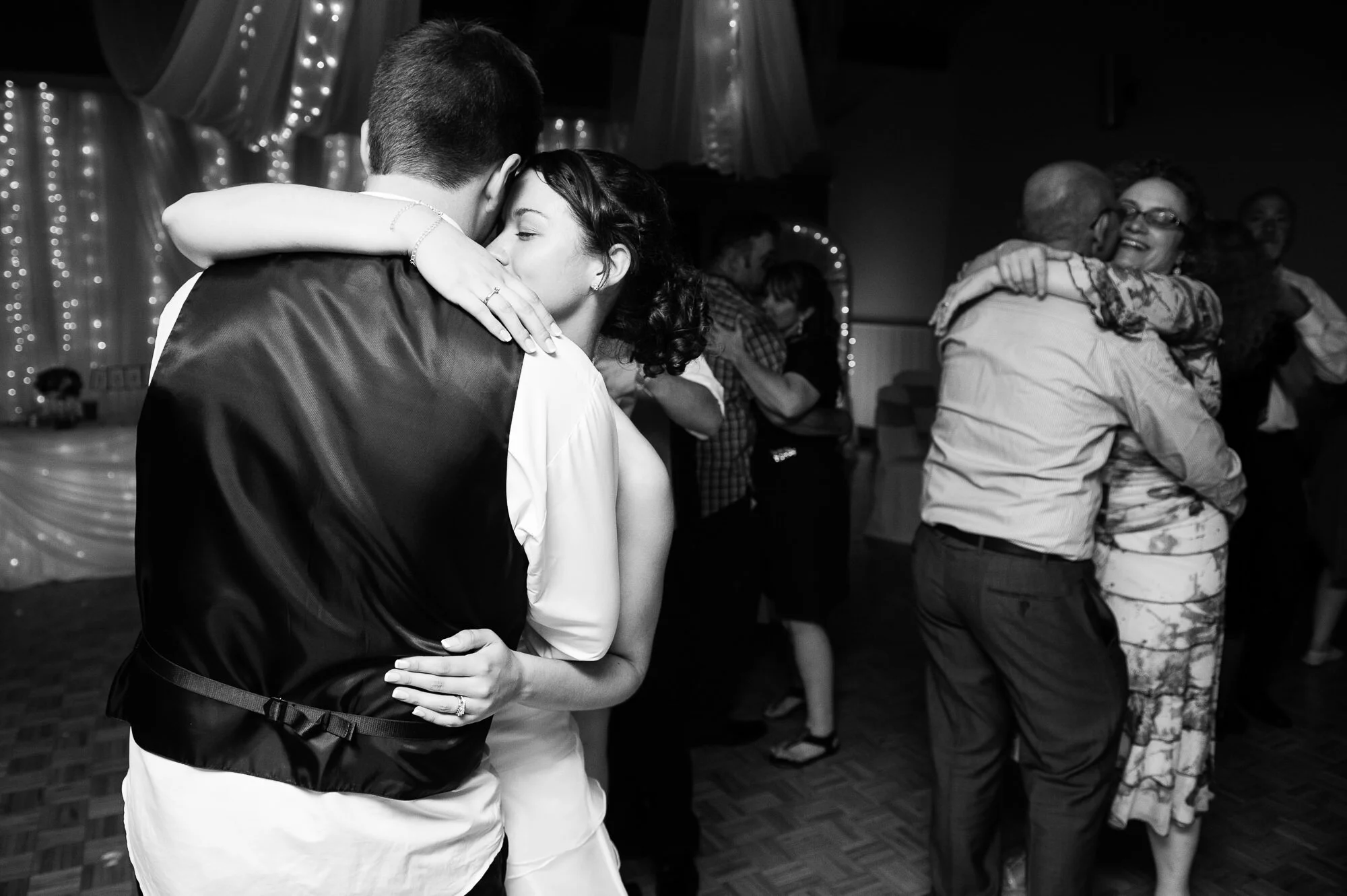 Couple dancing at a wedding reception with other couples in the background. Black and white photo with string lights and draped fabric as decor.