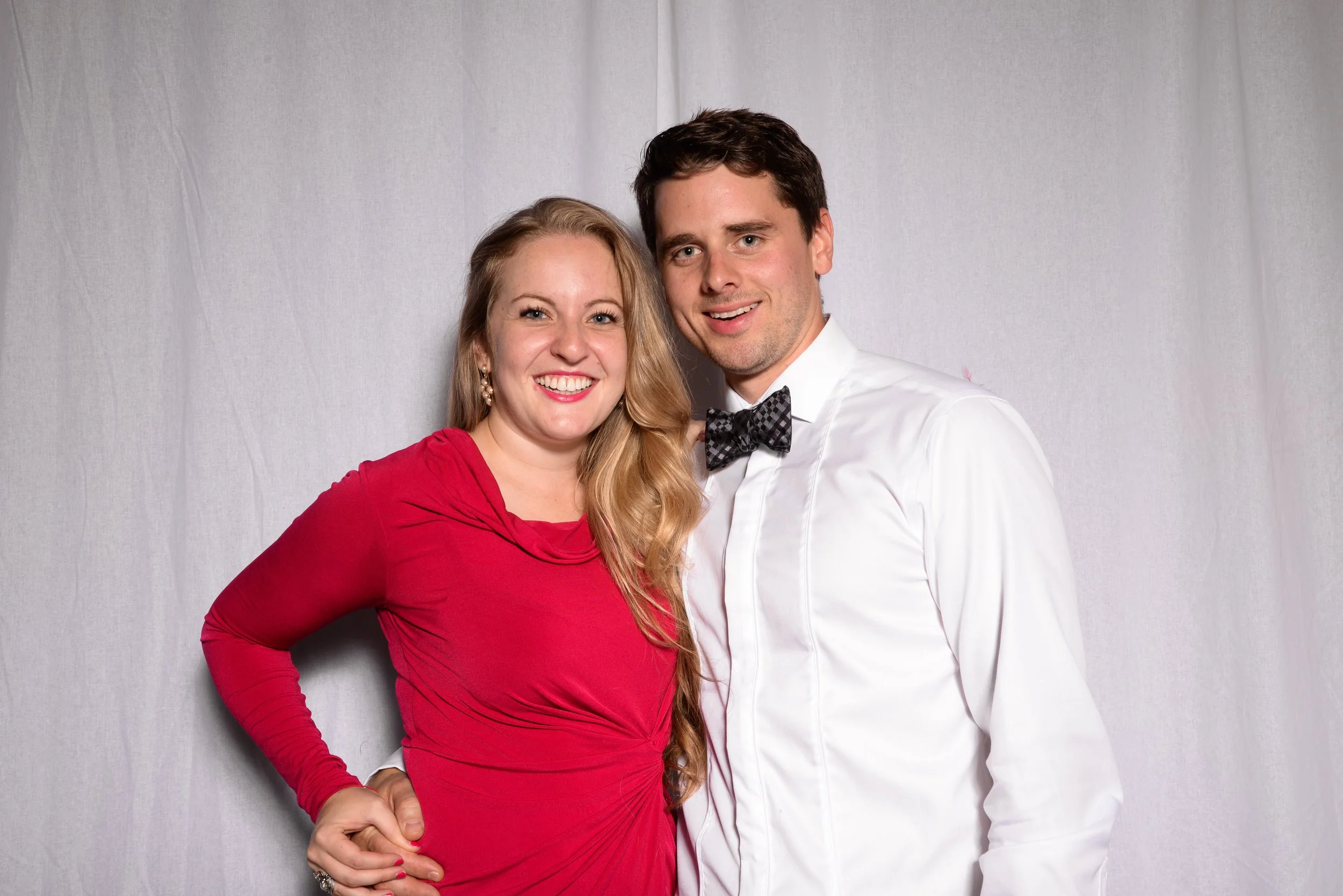 A smiling woman in a red dress and a man in a white shirt with a bow tie pose together against a white background.