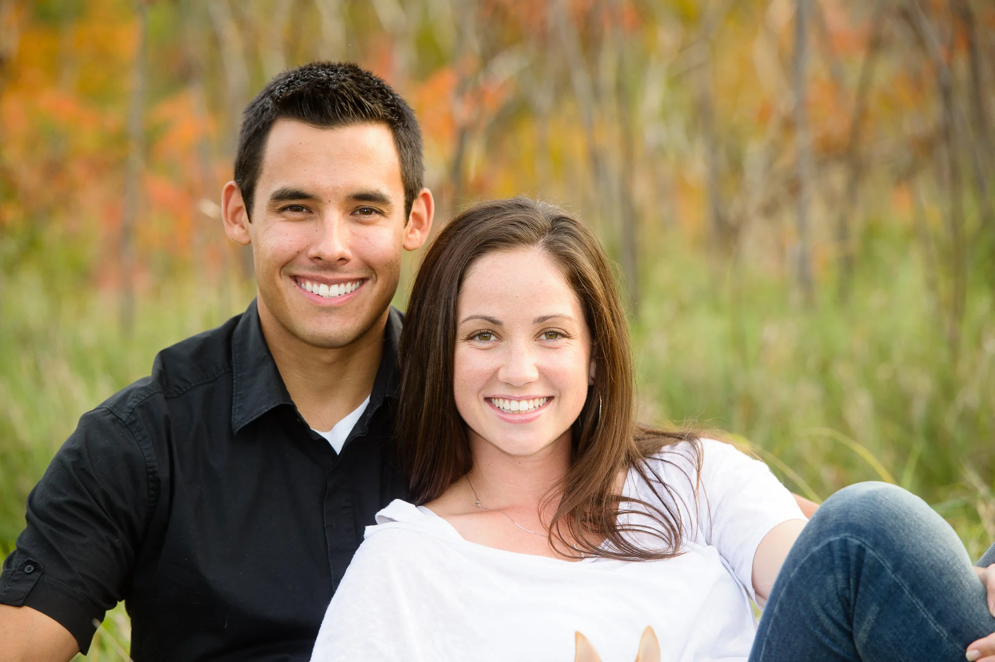 Smiling couple sitting outdoors in a grassy area with autumn foliage in the background.