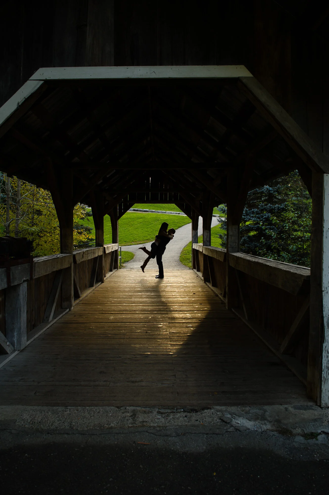Silhouette of a couple embracing on a wooden bridge with sunlight casting shadows, surrounded by greenery.