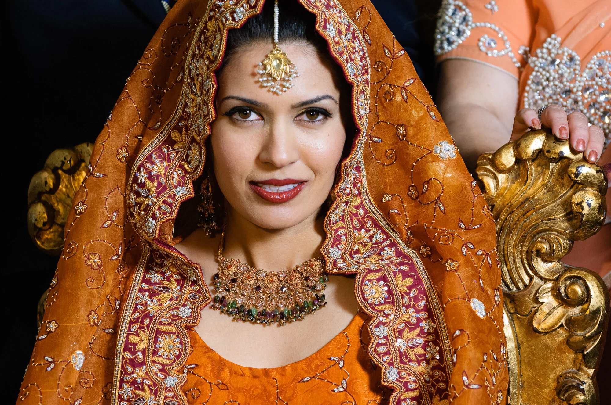 Woman in traditional orange embroidered attire with gold jewelry, sitting on ornate chair.