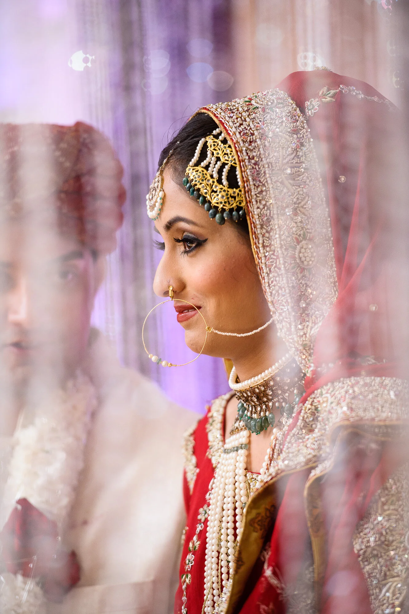 Bride in traditional South Asian wedding attire with ornate jewelry and headpiece, standing next to another person, partially visible, in ceremonial clothing.