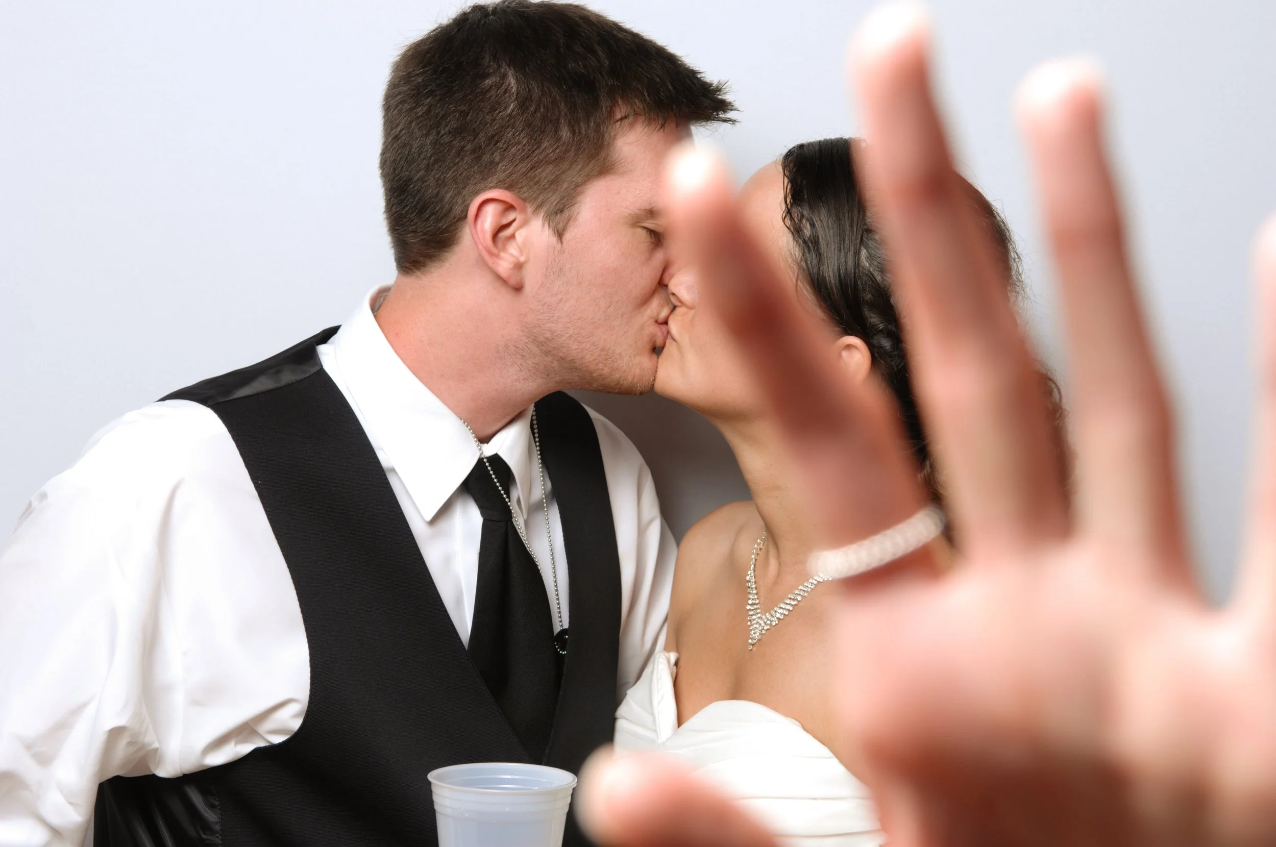 Wedding couple kissing with hand gesture in front, groom holding plastic cup, bride wearing pearl necklace.