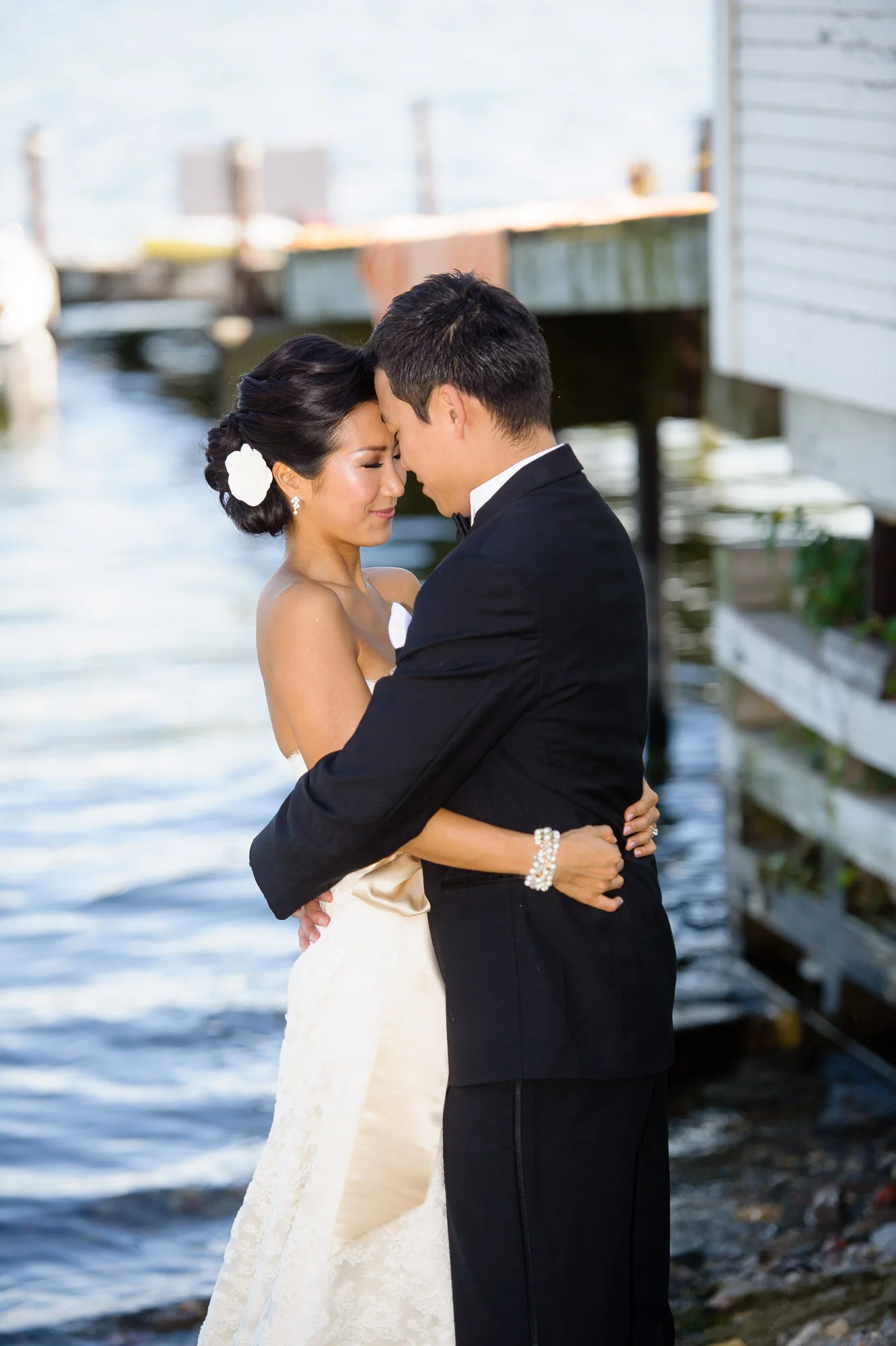 Bride and groom embracing near a lake, wearing formal wedding attire.