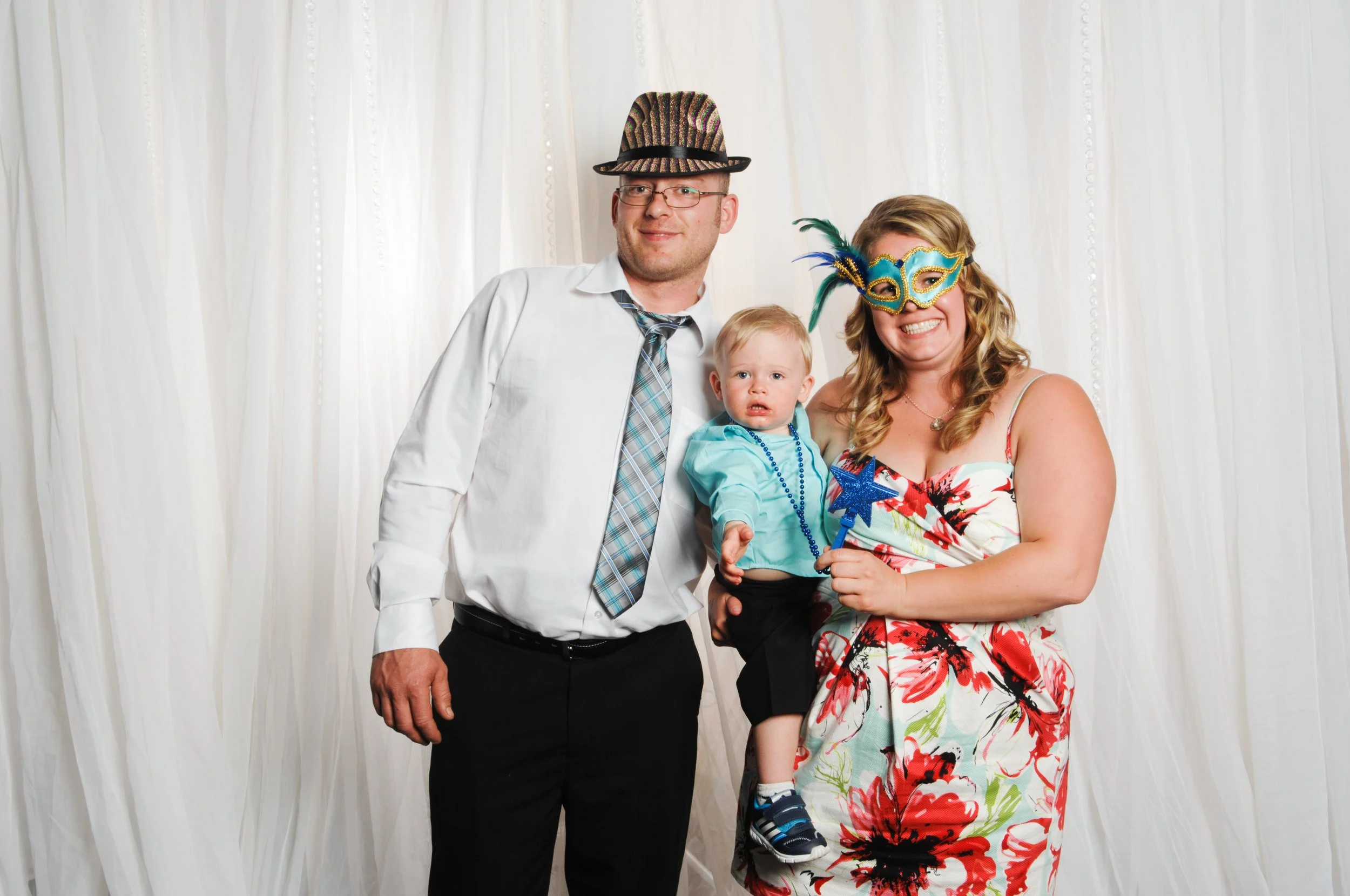 Family posing with hats and mask at a photo booth with white backdrop. Man in white shirt and tie, woman in floral dress, holding a child with blue shirt and beads.