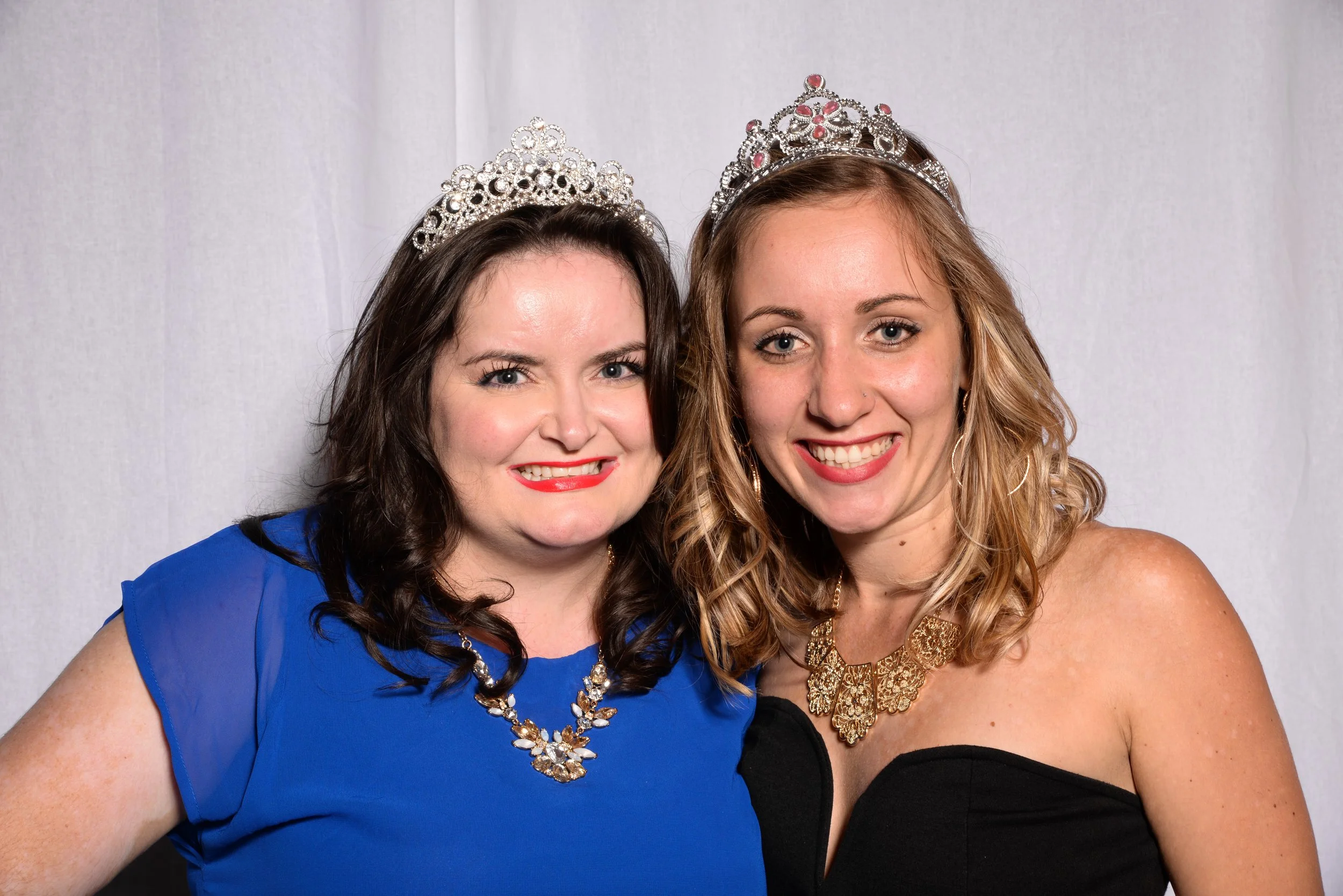 Two women wearing tiaras, smiling, one in a blue dress and the other in a black dress, against a white backdrop.